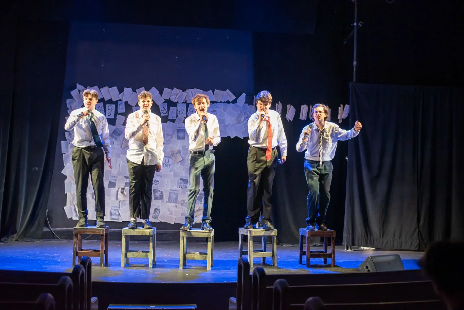 Five young boys dressed in white shirts, ties, and dark pants are performing on stage, standing on wooden chairs. They are holding microphones and appear to be singing or acting. The stage background features a display of papers and words, with curta