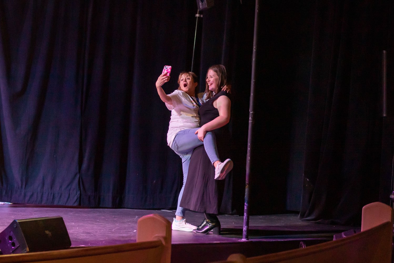 Two women are on stage, one holding a phone taking a selfie while the other woman is standing and smiling. The background shows black curtains and stage lighting.
