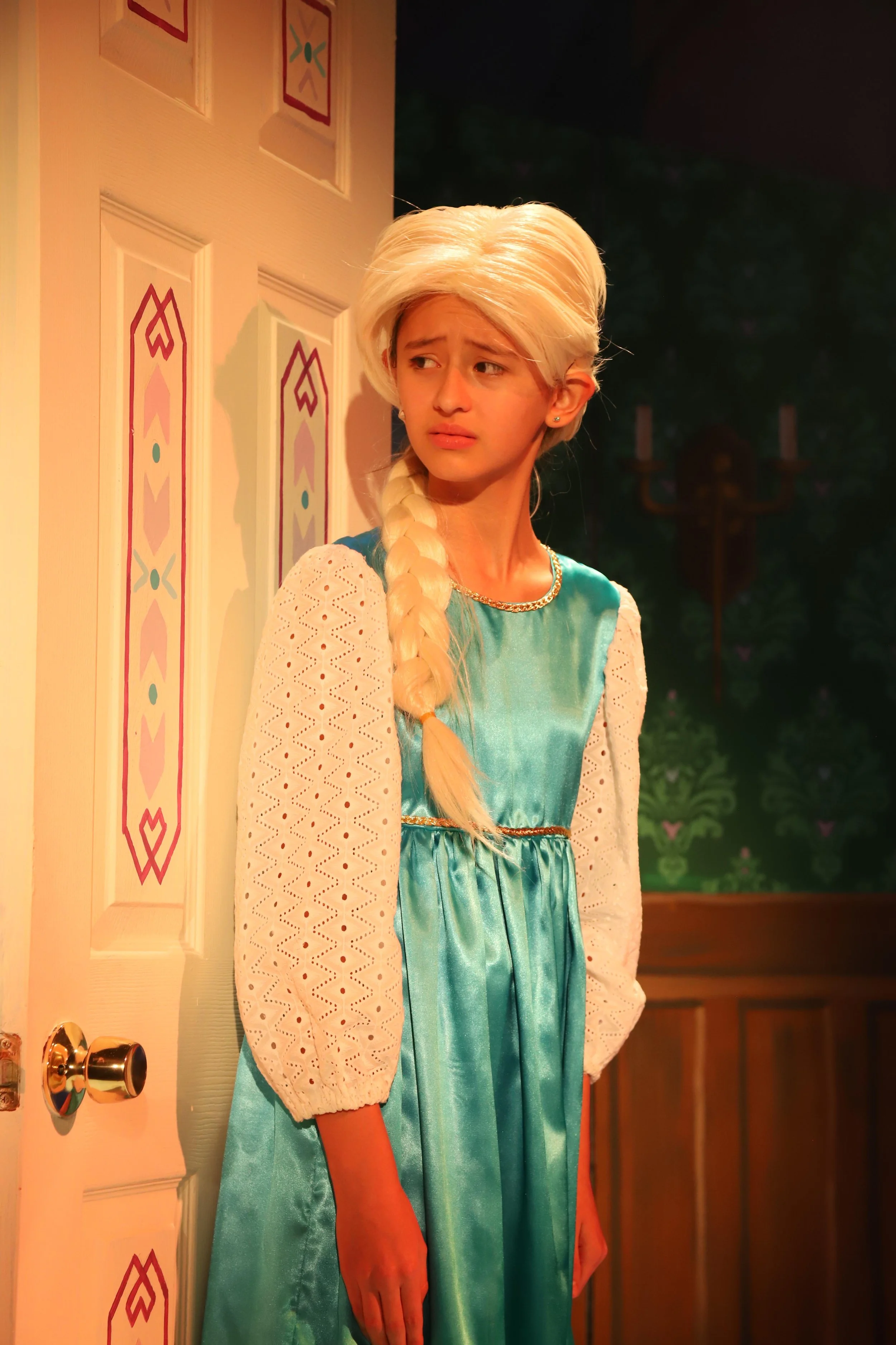 A young girl with long blonde hair in a braid, wearing a teal dress with pale eyes and a concerned expression, stands near a door with decorative panels, inside a warmly lit room.