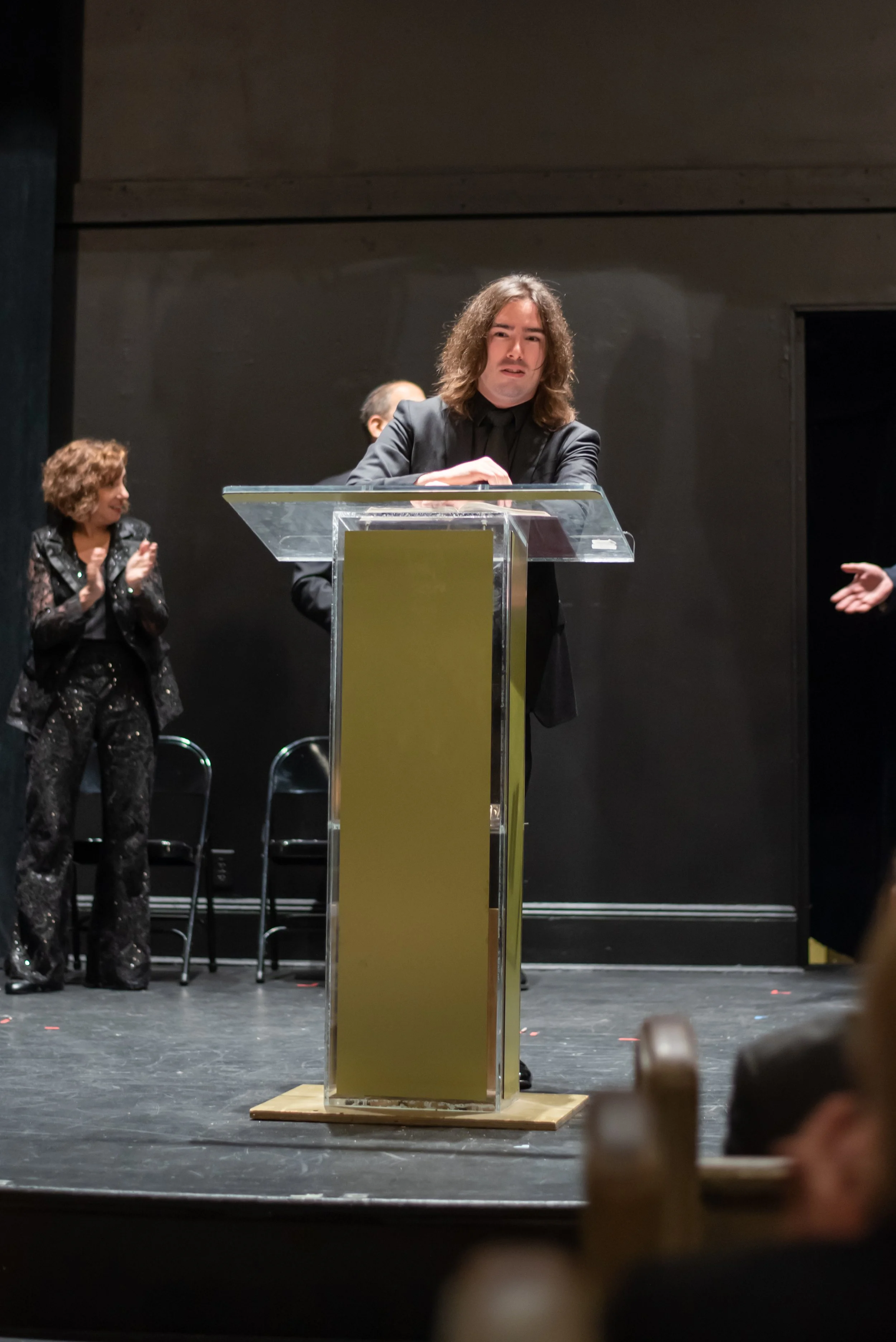 Man in black suit standing behind a clear podium in Grandma Sylvia's Funeral.  