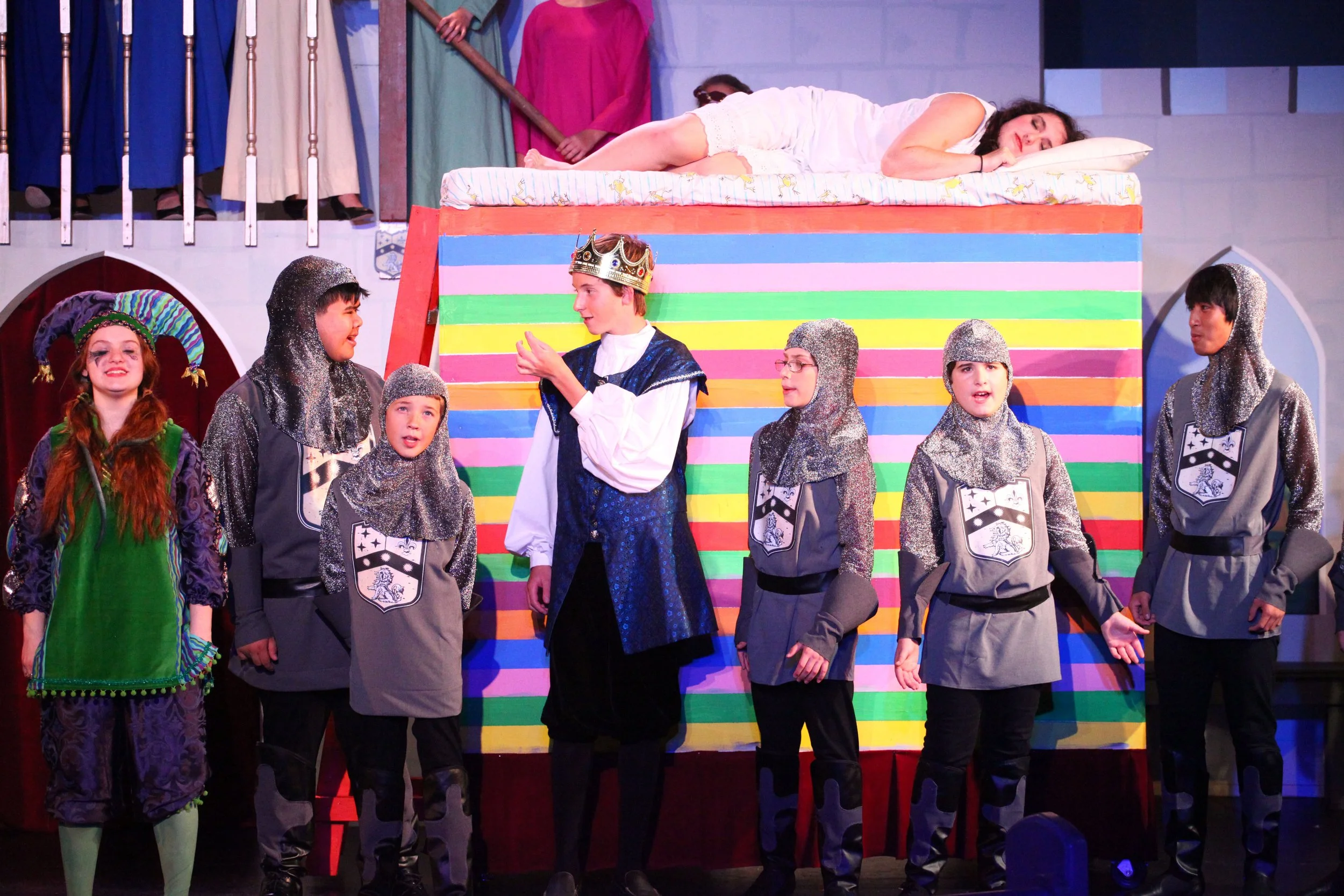 Children in costumes performing. One child in regal attire with a crown stands center, engaging with other children dressed in knight or medieval armor costumes. A girl in a colorful jester outfit smiles to the left. An adult woman lies on a bed abov