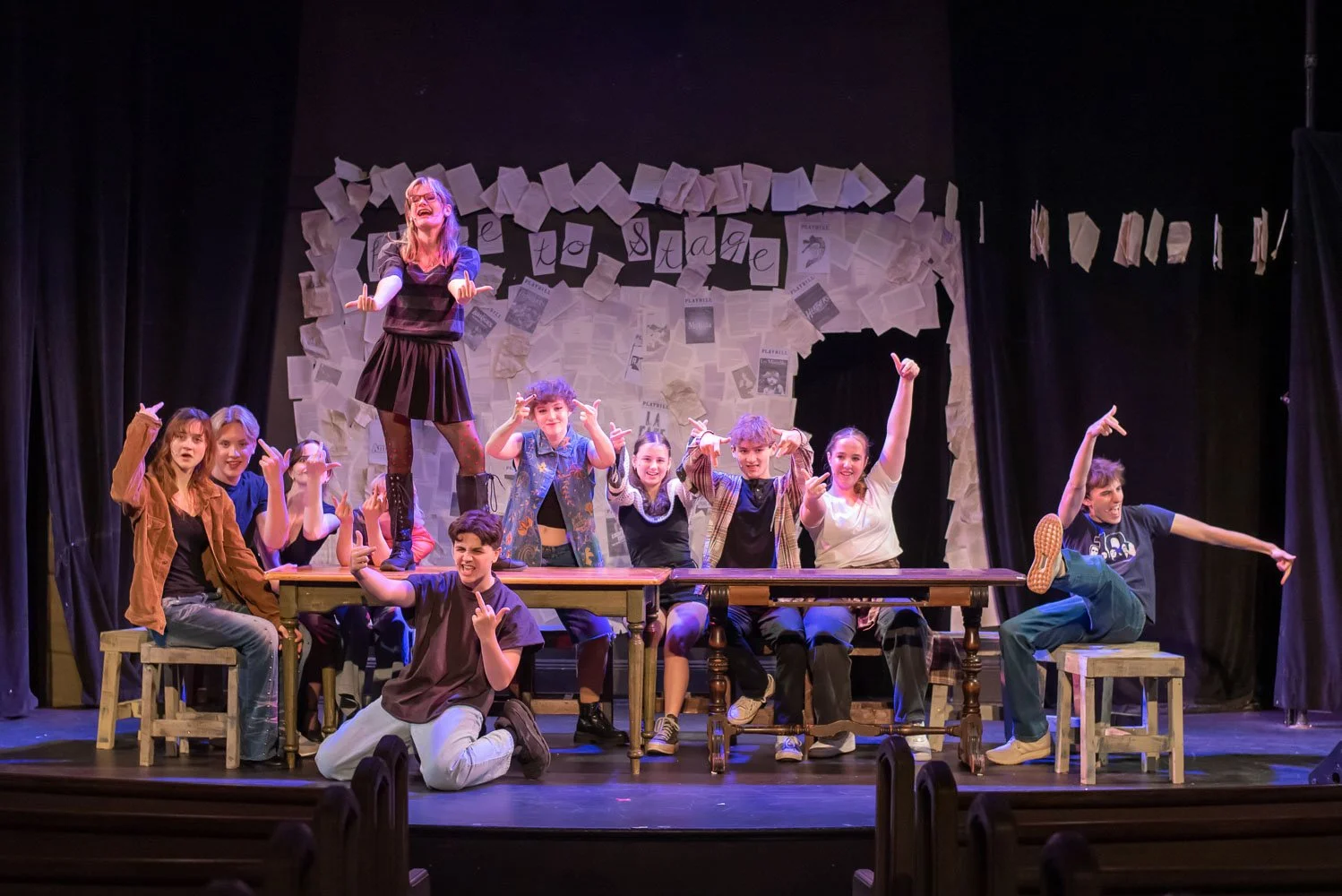 A group of children and a woman on stage during a theatrical performance, with a background decorated with papers and letters spelling 'Welcome to stage 2.' The woman, standing on a table, is engaging with the audience, while children around her make