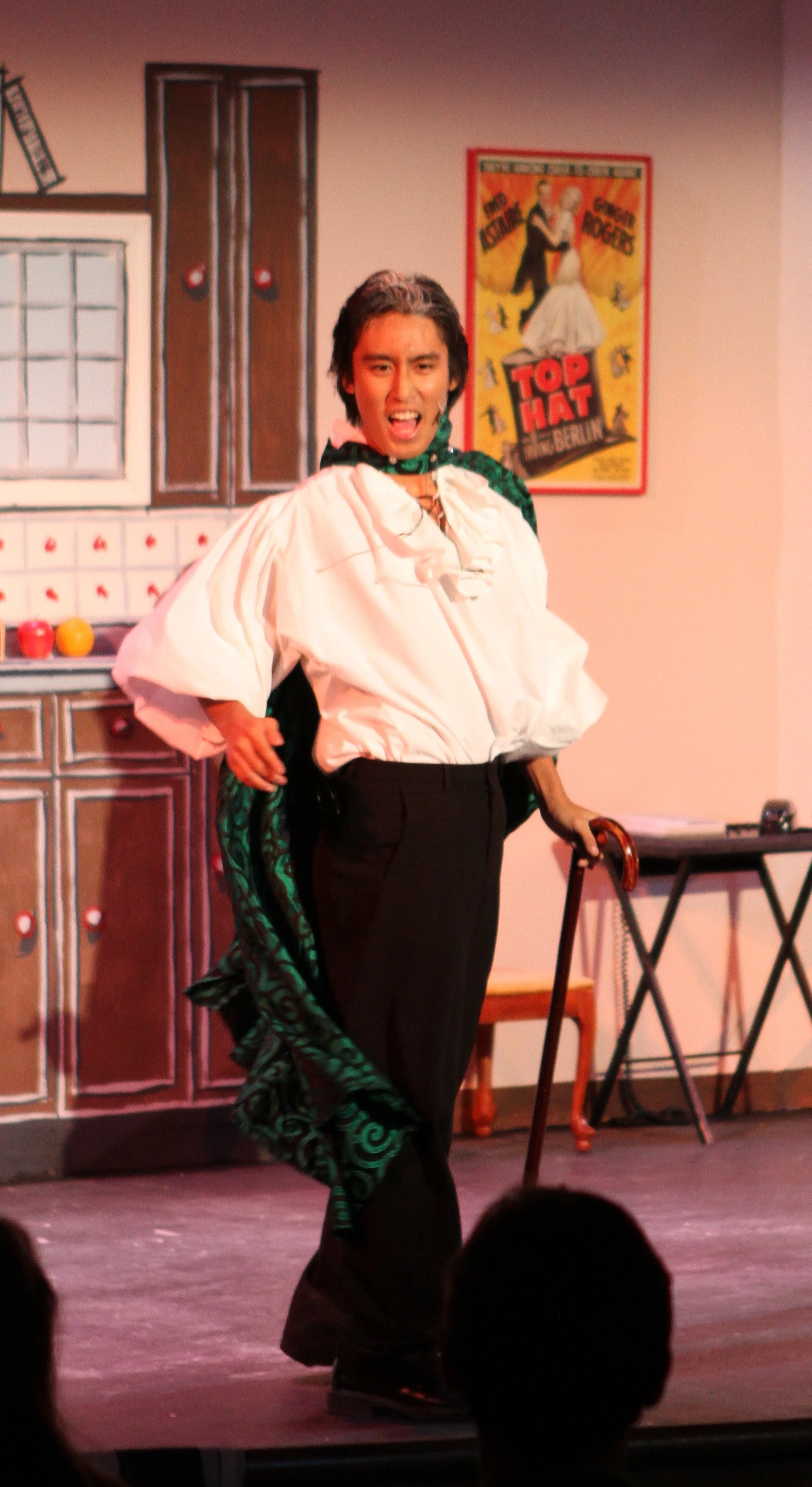 Performer on stage in Drowsy Chaperone, dressed in a white blouse, black pants, and a green scarf, holding a cane, with a backdrop resembling a kitchen and a poster that reads 'Top Hat'.