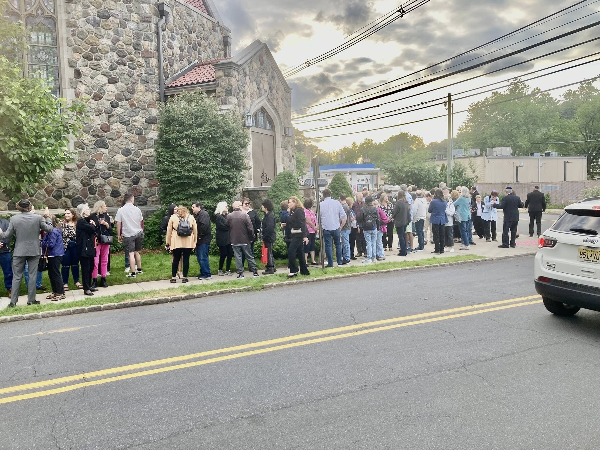 A large group of people is standing in a line outside a building with stone walls, on a sidewalk next to a street with yellow dividing lines, in the early evening or late afternoon. Scene from the play Grandma Sylvia's Funeral.