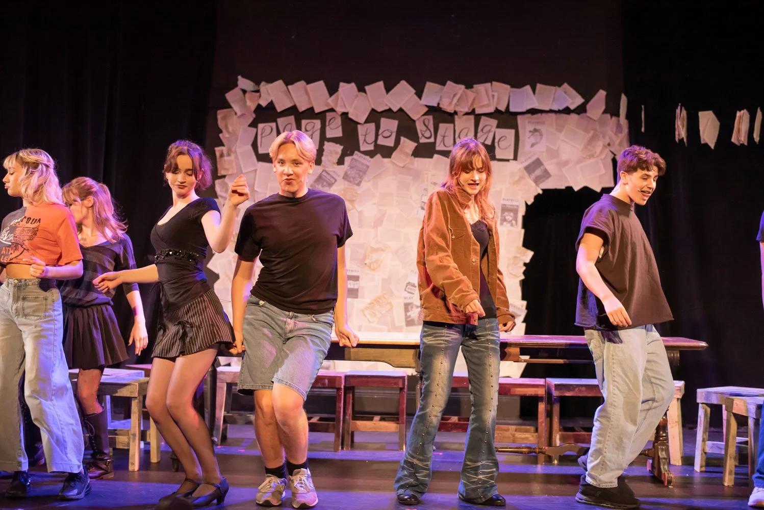 Teenagers performing a dance or scene on stage with a backdrop of newspaper clippings and paper signs. Performance of Page to Stage cabaret.