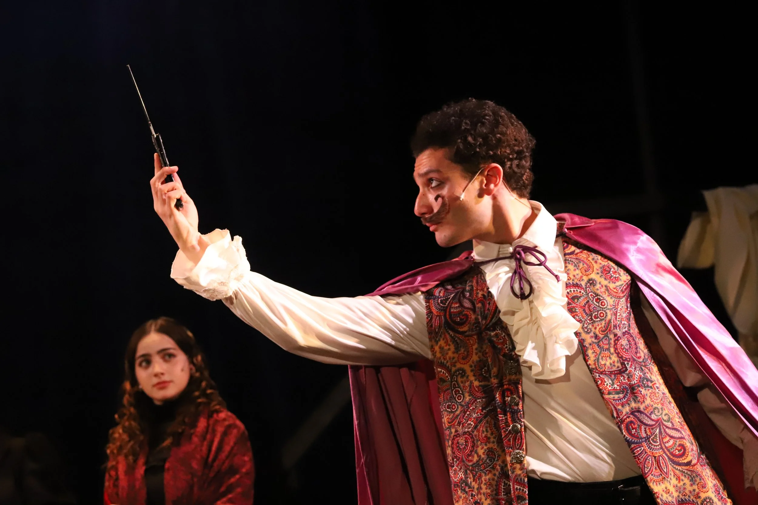 A man dressed as a nobleman on stage, holding a sword, with a woman in a red costume watching him in the background.