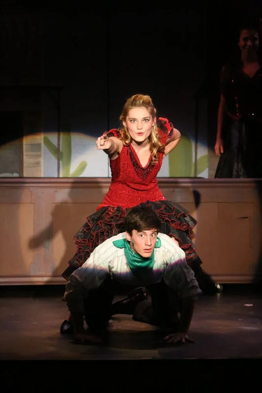 A woman in a red dress is sitting on the back of a man in a white shirt and green bandana, who is on all fours. She is pointing forward with a confident expression, while he looks surprised. There is a person in black in the background on a stage.