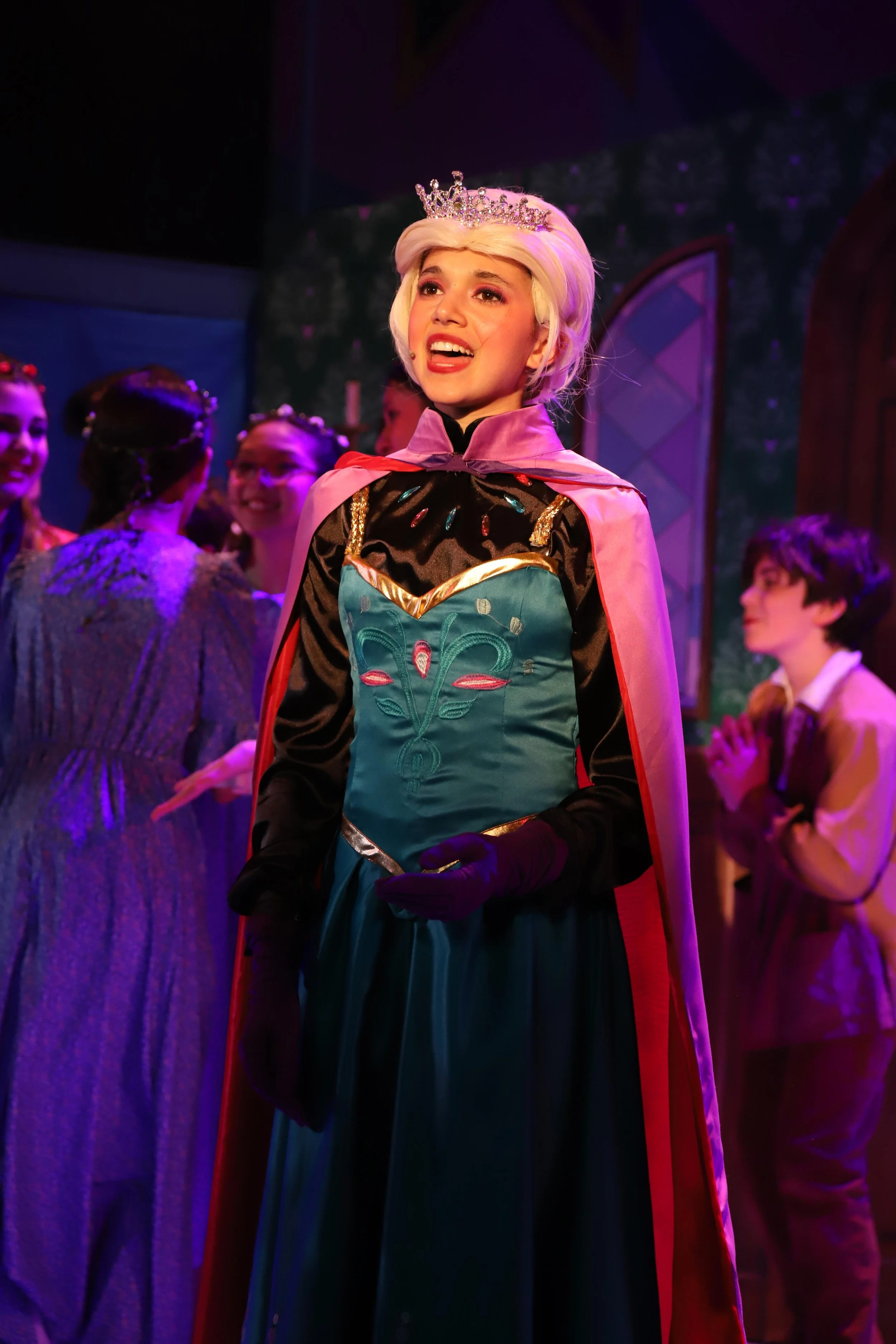 Performers on stage, with a woman in an elaborate costume with a crown and cape, singing or speaking. Women in dresses and children are visible in the background.