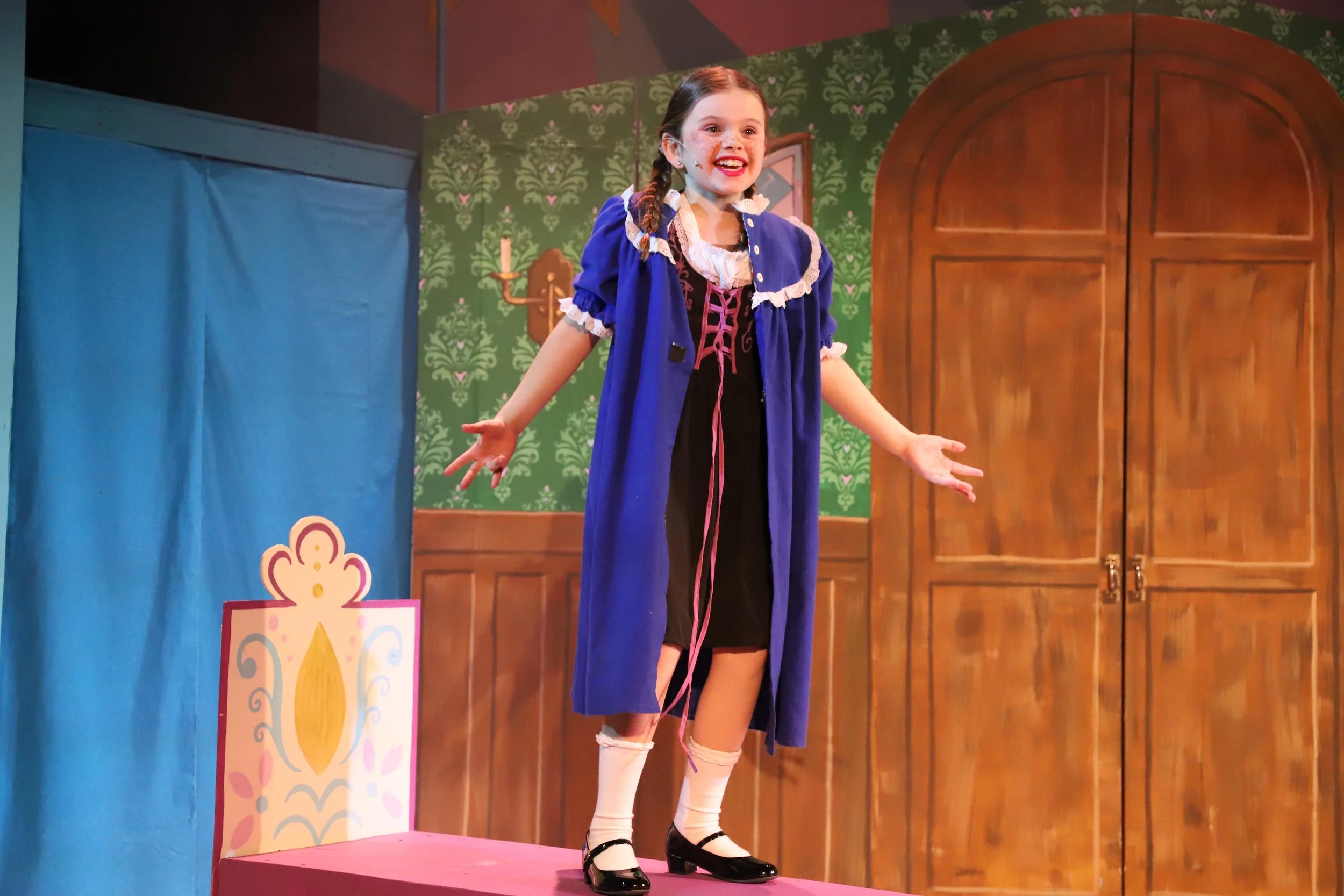 Young girl in a colorful costume performing on stage in a theater production of Frozen Jr. with a backdrop of a green patterned wall and a wooden door, and a pink and blue stage set.
