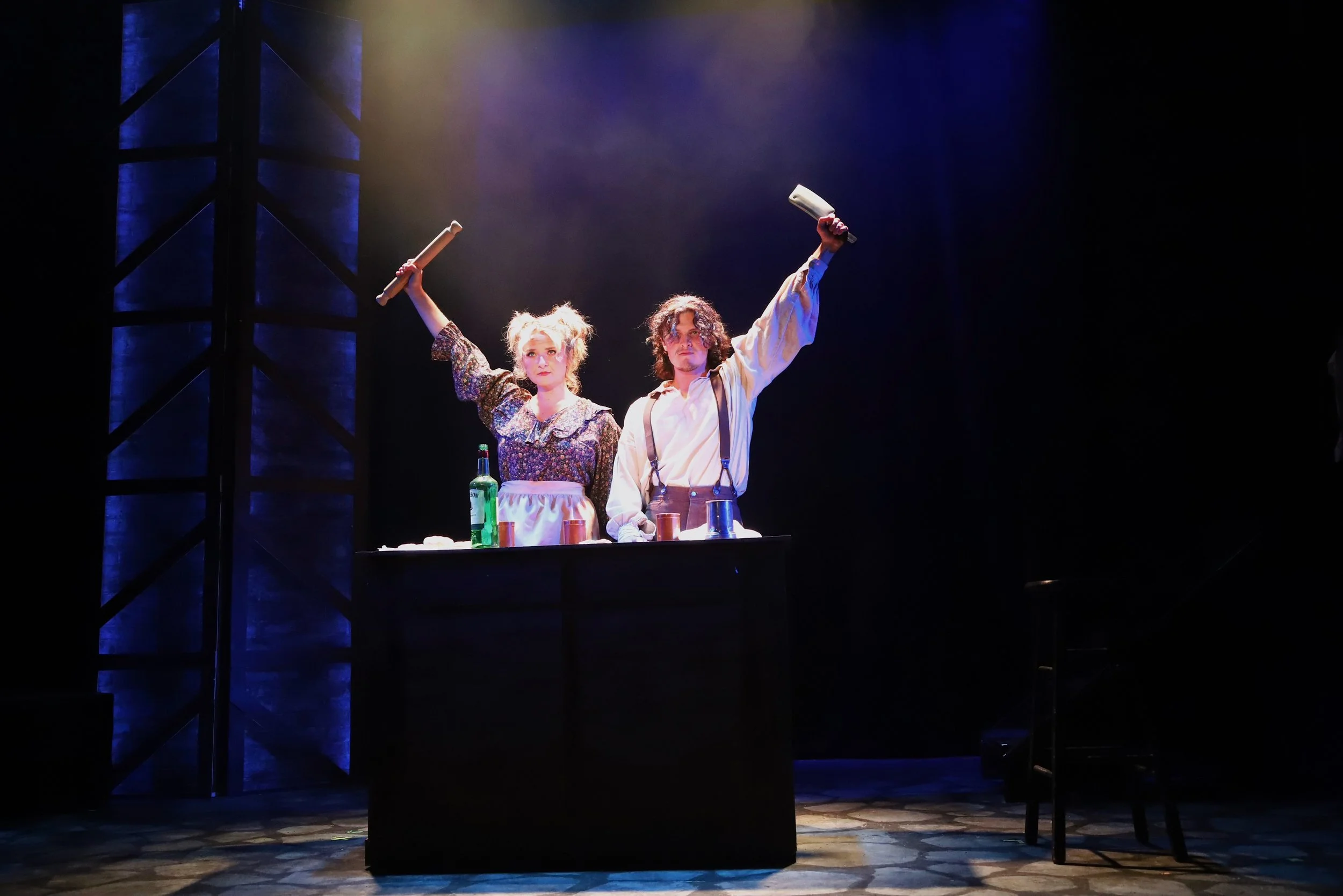 Two actors on stage, one woman and one man, standing behind a black table with bottles and glasses, raising rolling pins in the air, illuminated by stage lighting with a dark background.