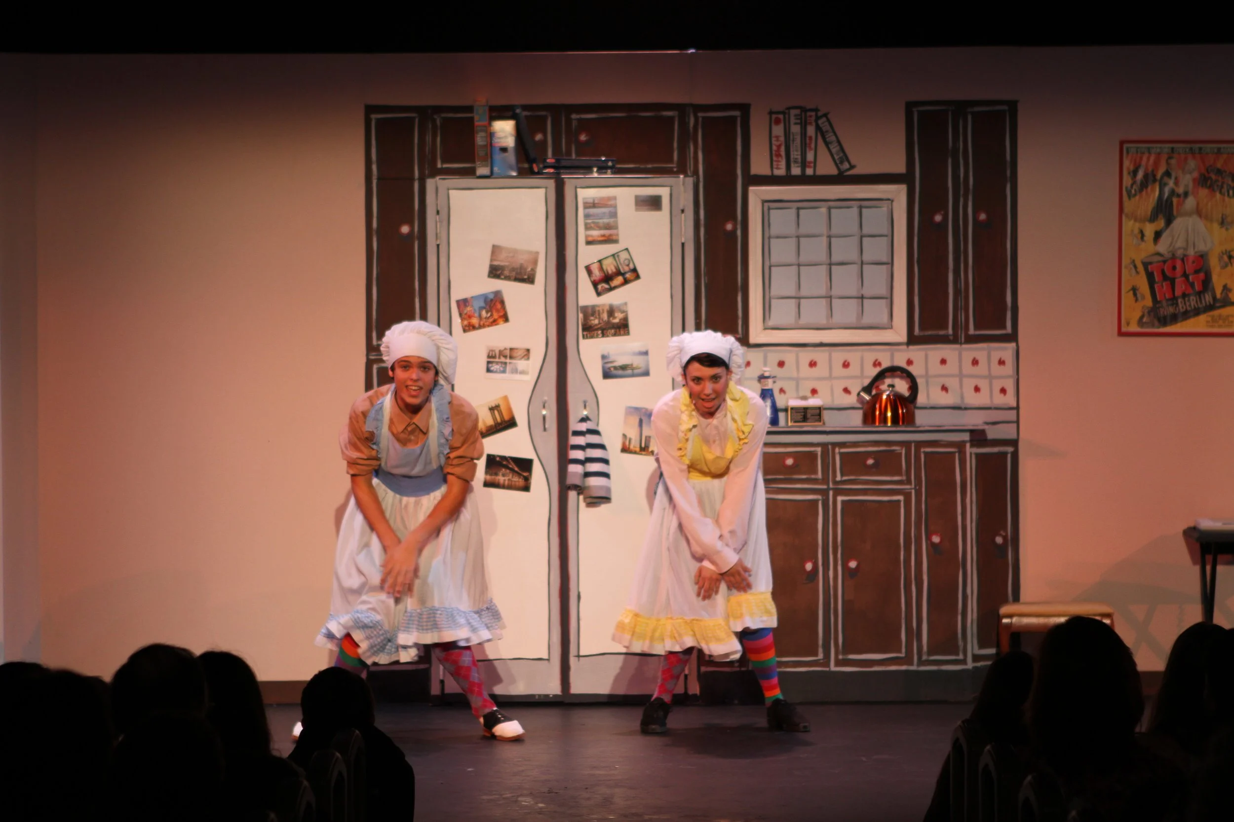 Two performers on stage in Drowsy Chaperone in colorful costumes, dancing in front of a painted kitchen set with a fridge, cupboards, and a poster on the wall.