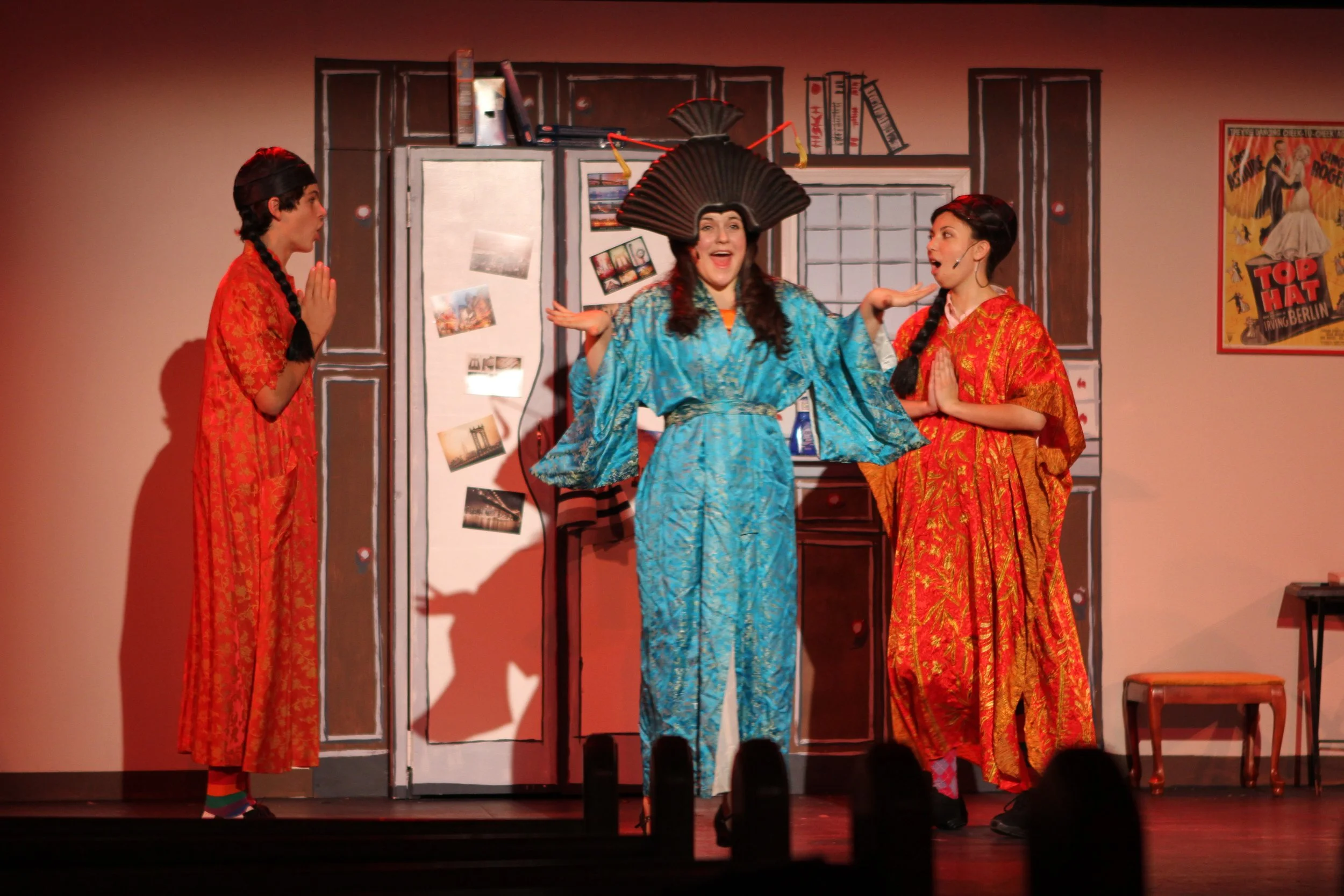 Three women on stage in colorful costumes, with the middle woman wearing a large black fan hat and a blue silk robe, appearing to perform a comedic scene. The stage background has a painted door and wall decor, including a movie poster on the right.