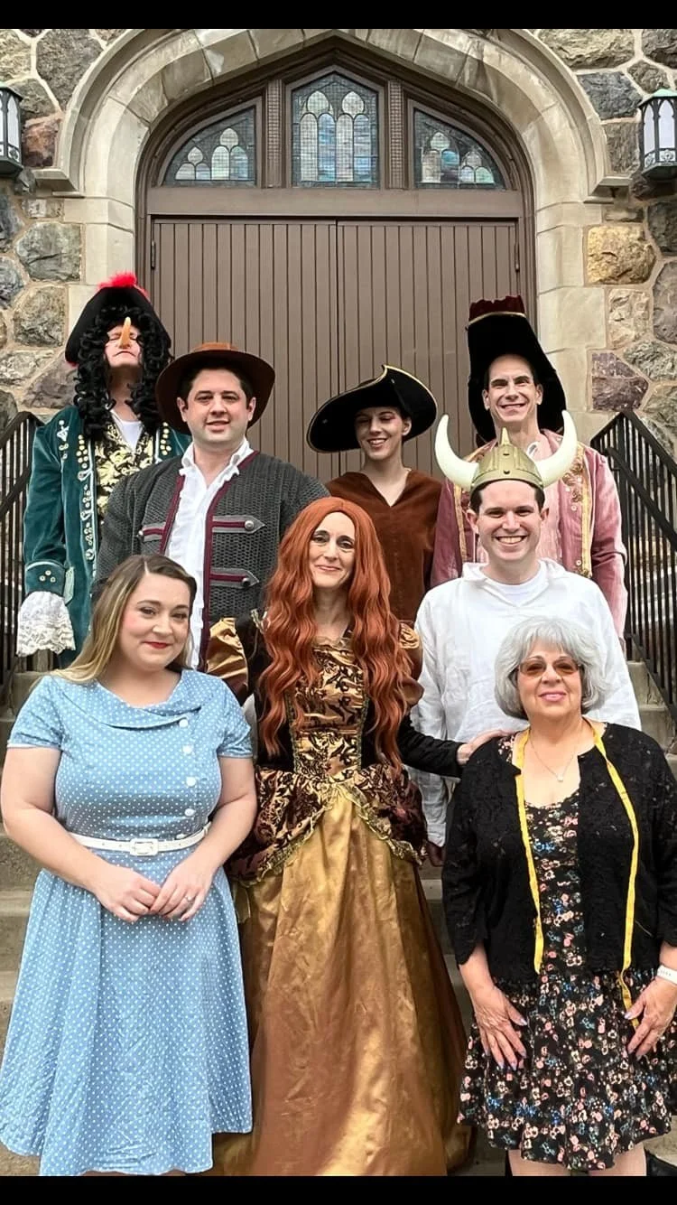 Group of people dressed in costumes of well-known characters from 'The Lord of the Rings' and 'The Hobbit' standing on stone stairs in front of a wooden door with stained glass above.