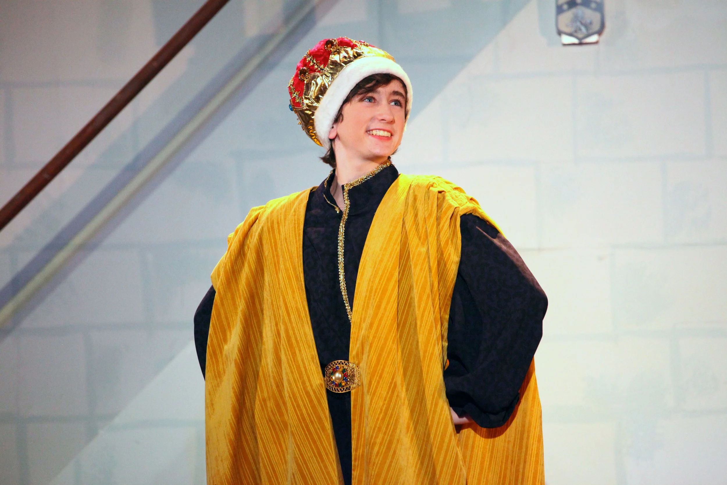 A person dressed in medieval royal attire, wearing a crown and a yellow cloak, smiling and standing indoors. Scene from Once Upon a Mattress.