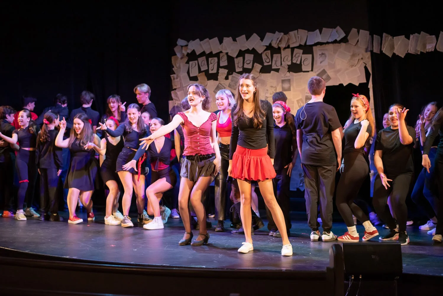 A group of young performers on stage during a theatrical performance, with some dancing and smiling, background decorated with papers and letters spelling 'Page to Stage'.