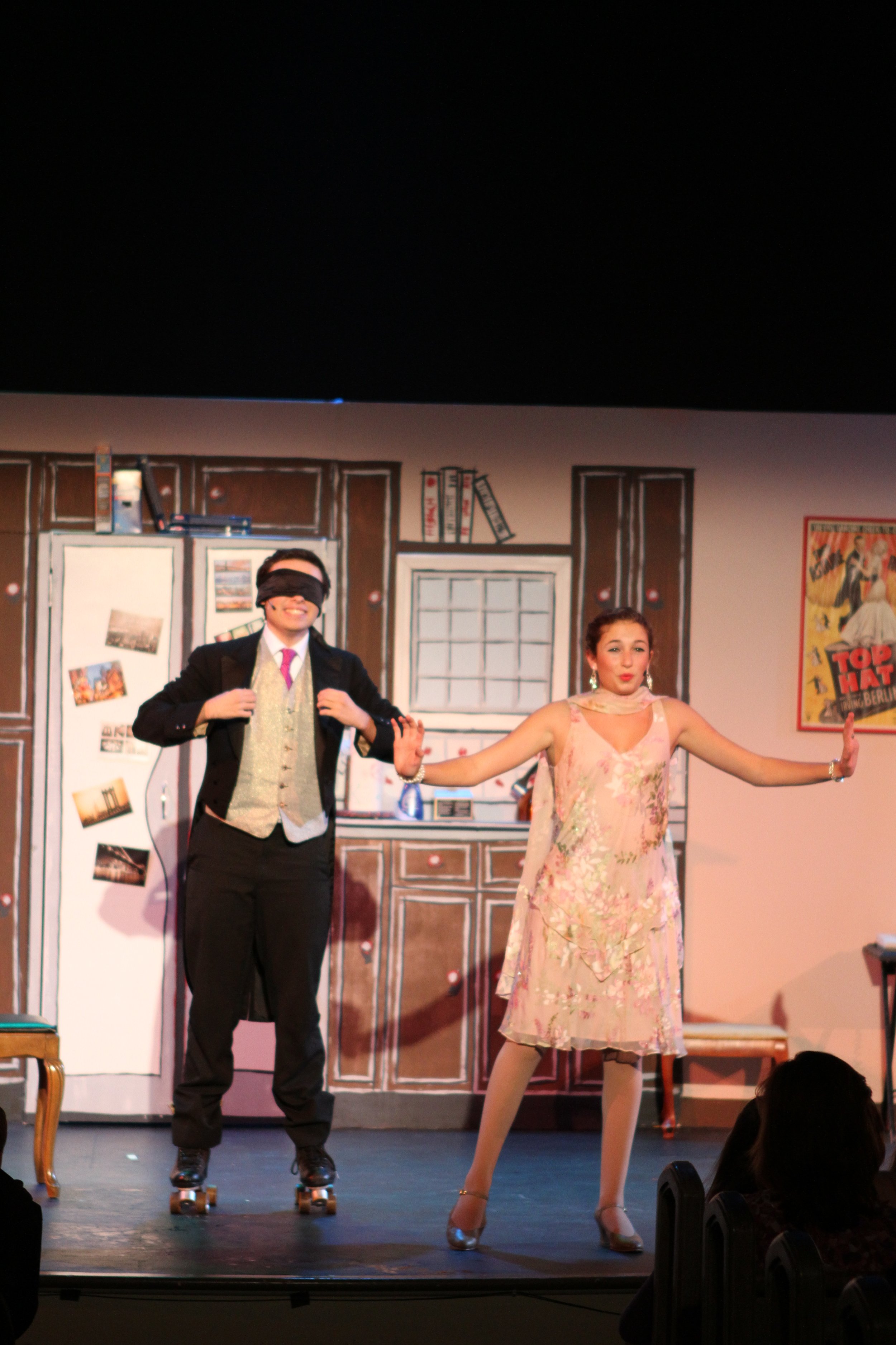 Two performers on stage, one man with a black blindfold, dressed in a black suit with pink tie and gray vest, holding hands with a woman in a pink floral dress, with a painted kitchen scene in the background.