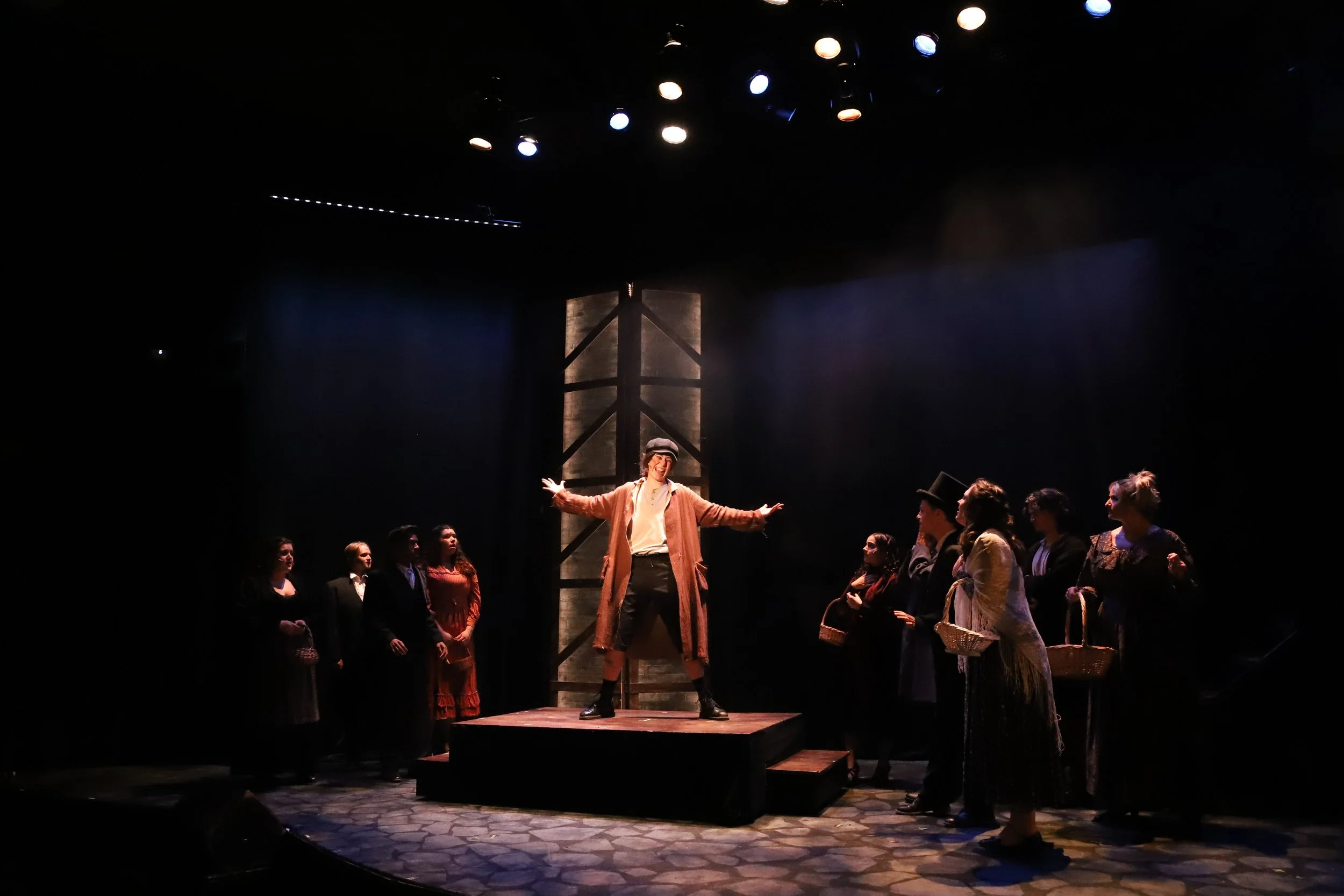 Theater scene with actor in center on a small stage, surrounded by other actors in vintage costumes, under stage lighting.