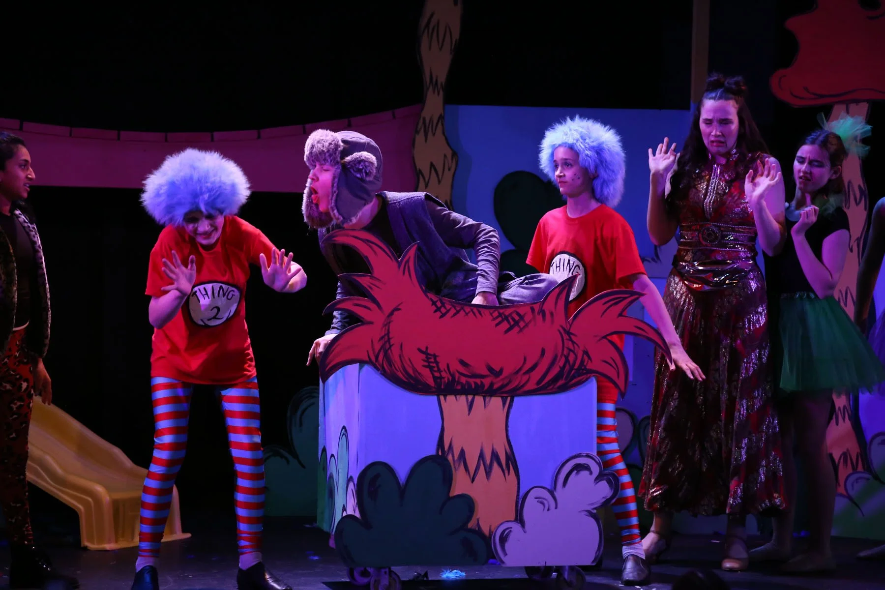 Children performing a play on stage dressed in colorful costumes, including wigs and striped tights, with a tree and cartoon scenery painted backdrop. Scene from Seussical Jr.  