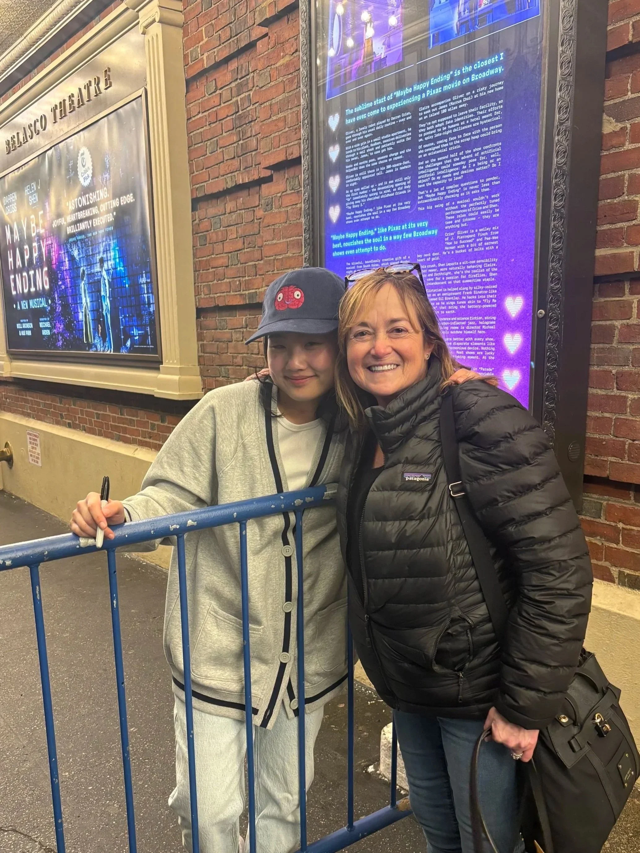 Helen J. Shen and Jayne Myers smiling behind a blue barricade outside Belasco Theatre, with illuminated posters and theater marquee in the background. Show is Maybe Happy Ending.