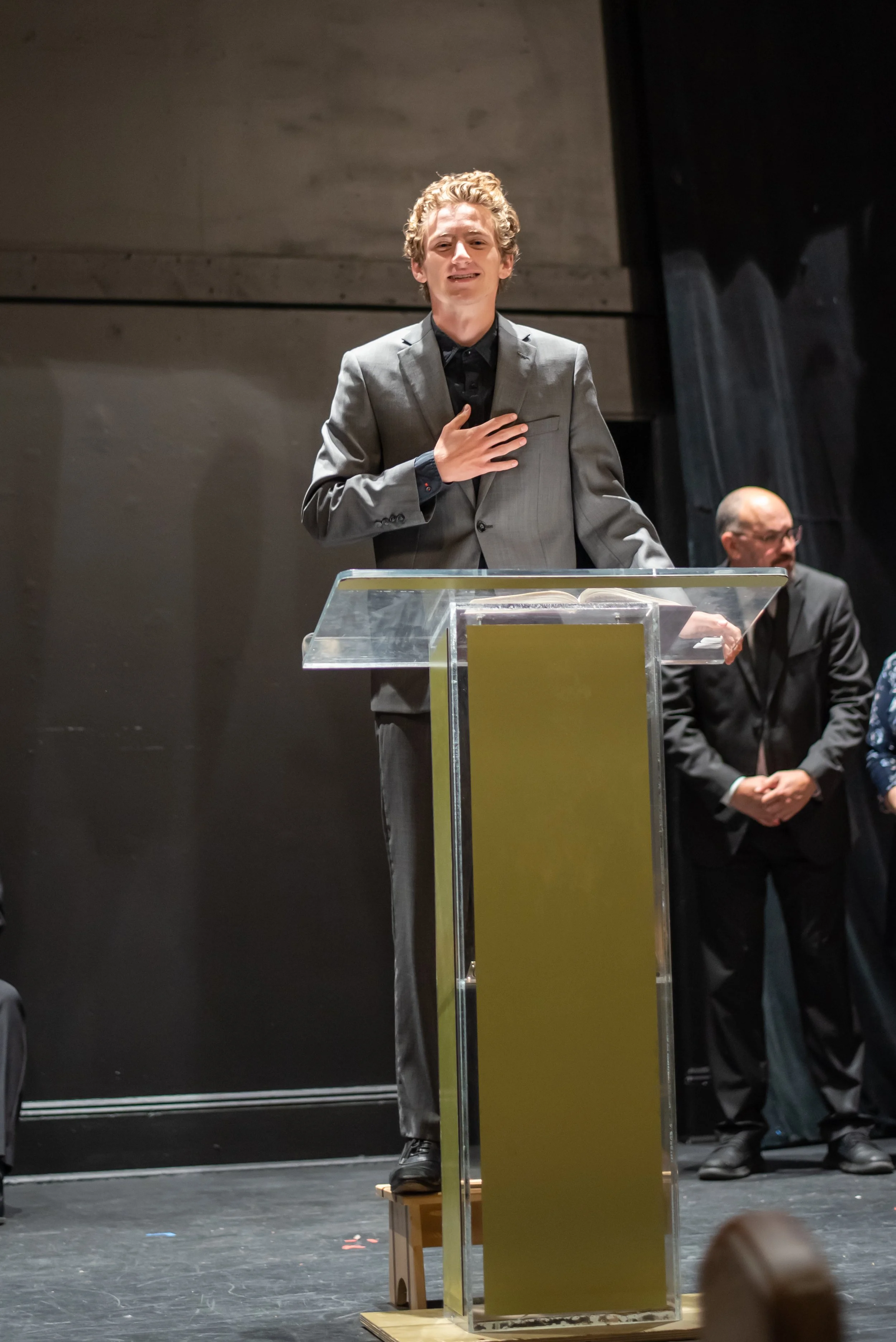 A young man in a gray suit stands on a small stool at a podium, with his right hand over his heart and his mouth slightly open, in a scene from Grandma Sylvia's Funeral. Other individuals stand behind him.