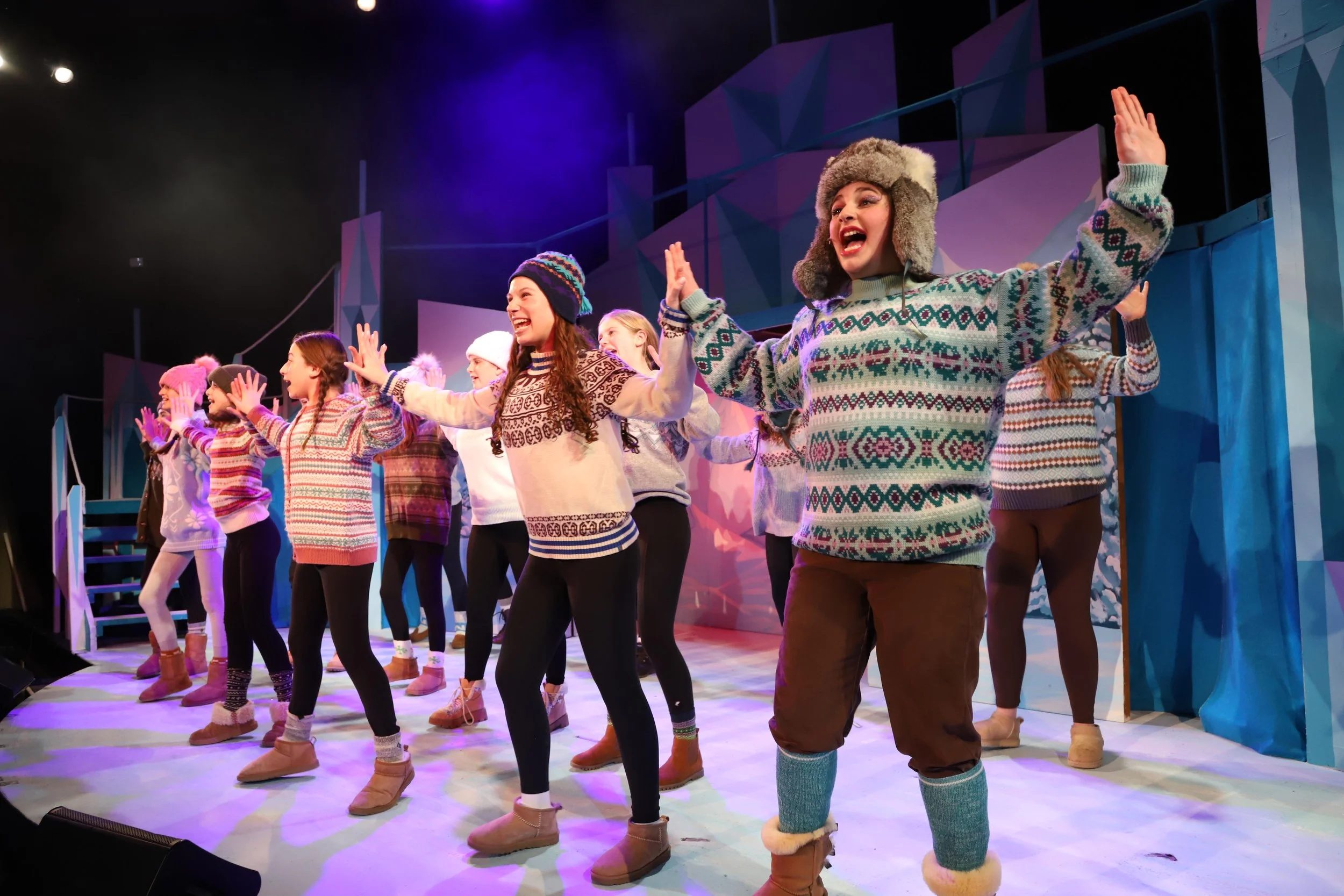 Group of women performing on stage, wearing winter sweaters, hats, and boots, with a colorful backdrop and stage lighting.