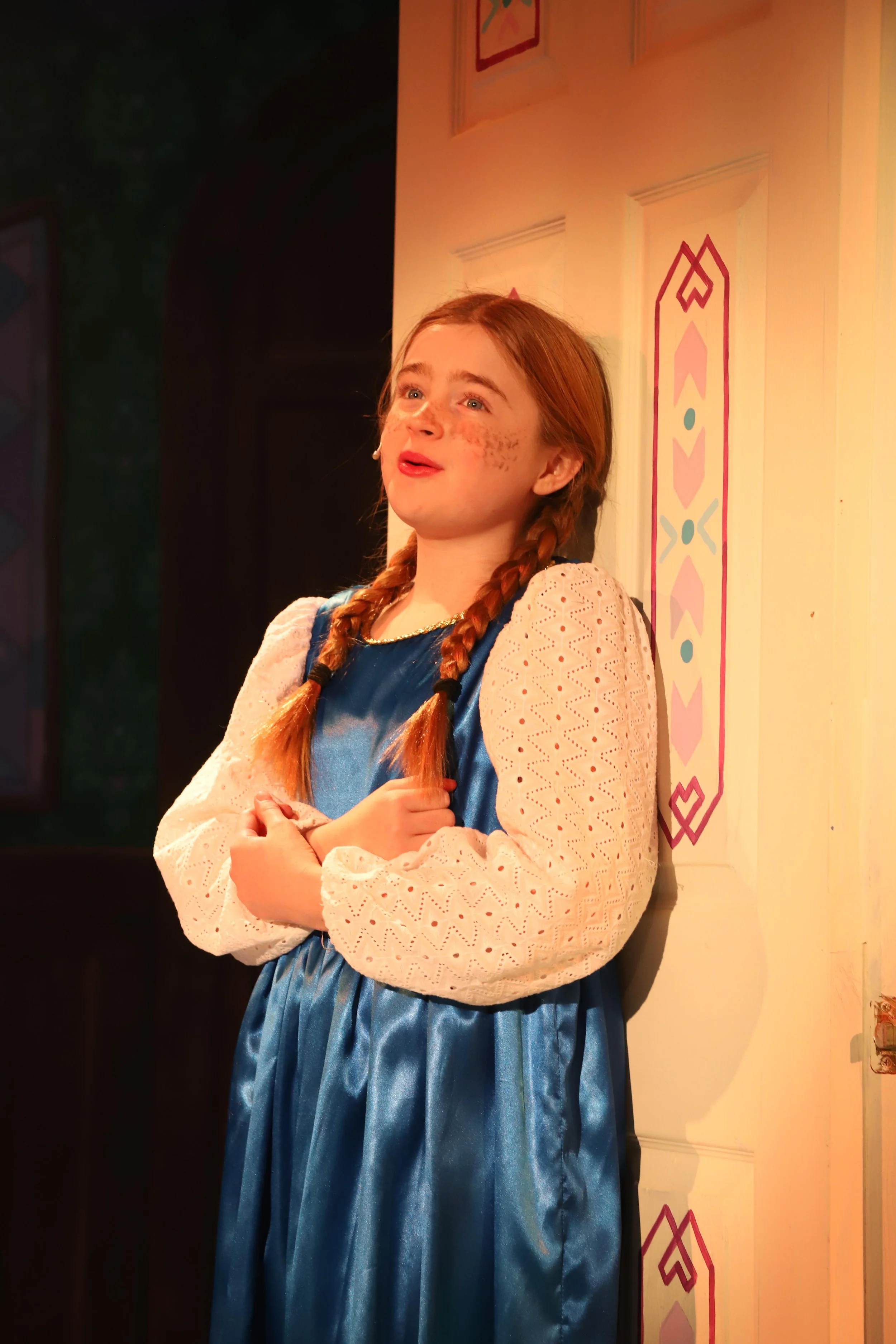 A young girl with red hair in braids, wearing a blue dress with white lace sleeves, standing on stage with a painted backdrop.