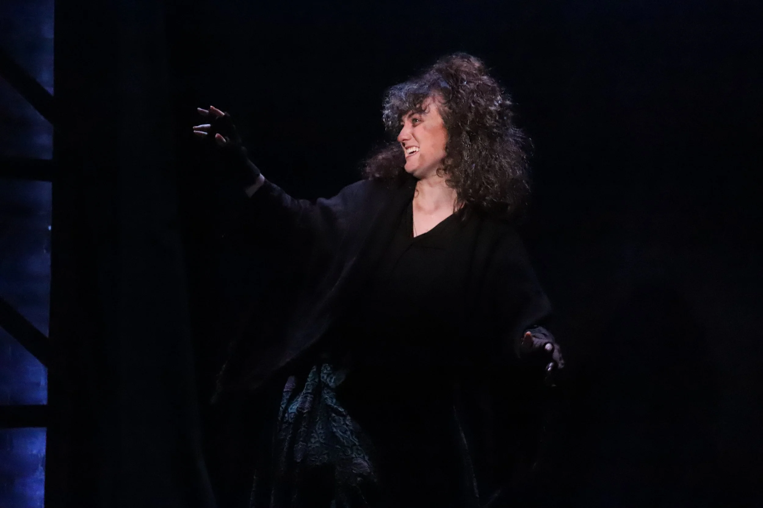 A woman with curly hair and a black outfit laughing and gesturing with her right hand in a dark setting.