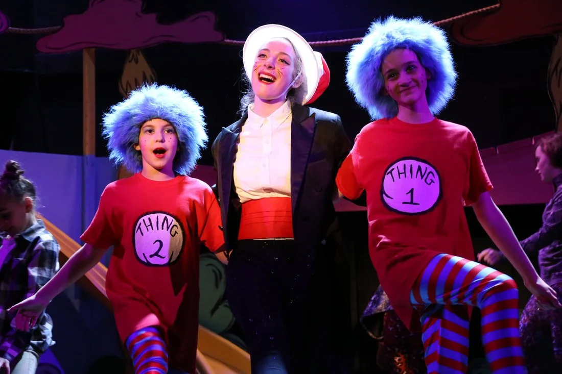 Three performers on stage dressed as characters from 'The Cat in the Hat,' with the one in the middle smiling and wearing a tall white hat, flanked by two children with blue fuzzy hats and red shirts labeled 'Thing 1' and 'Thing 2'.