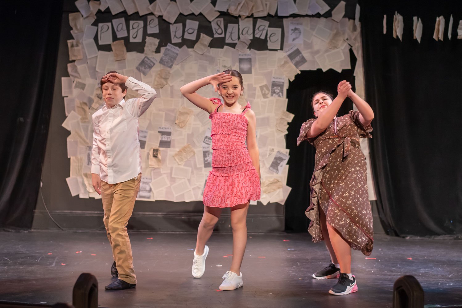 Three children performing a stage play, standing with hand gestures, in front of a backdrop decorated with torn paper and photographs. Performance of Page to Stage cabaret.