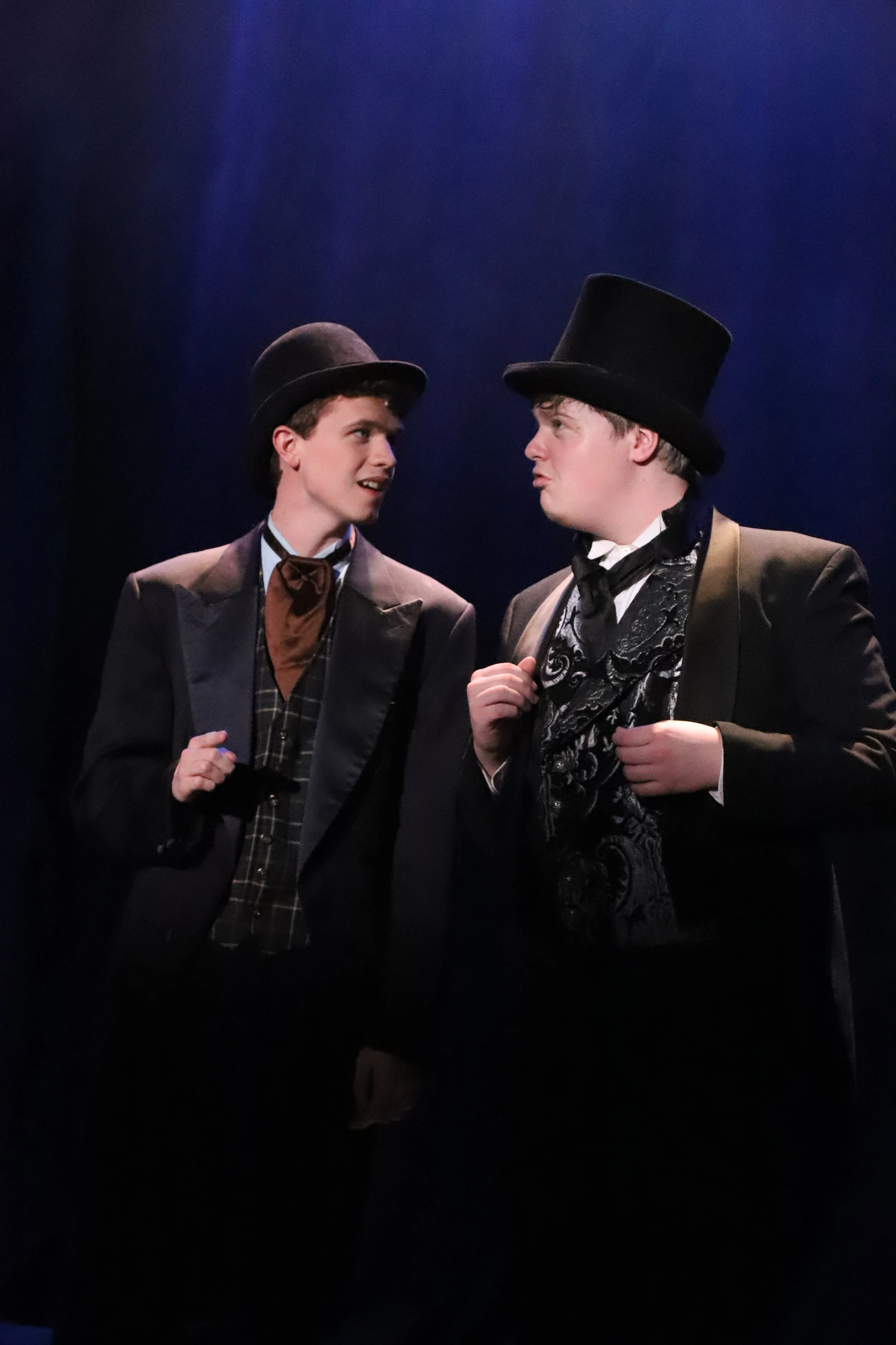 Two young men in 19th-century costumes engaging in conversation on stage, one adjusting his waistcoat, both wearing top hats.