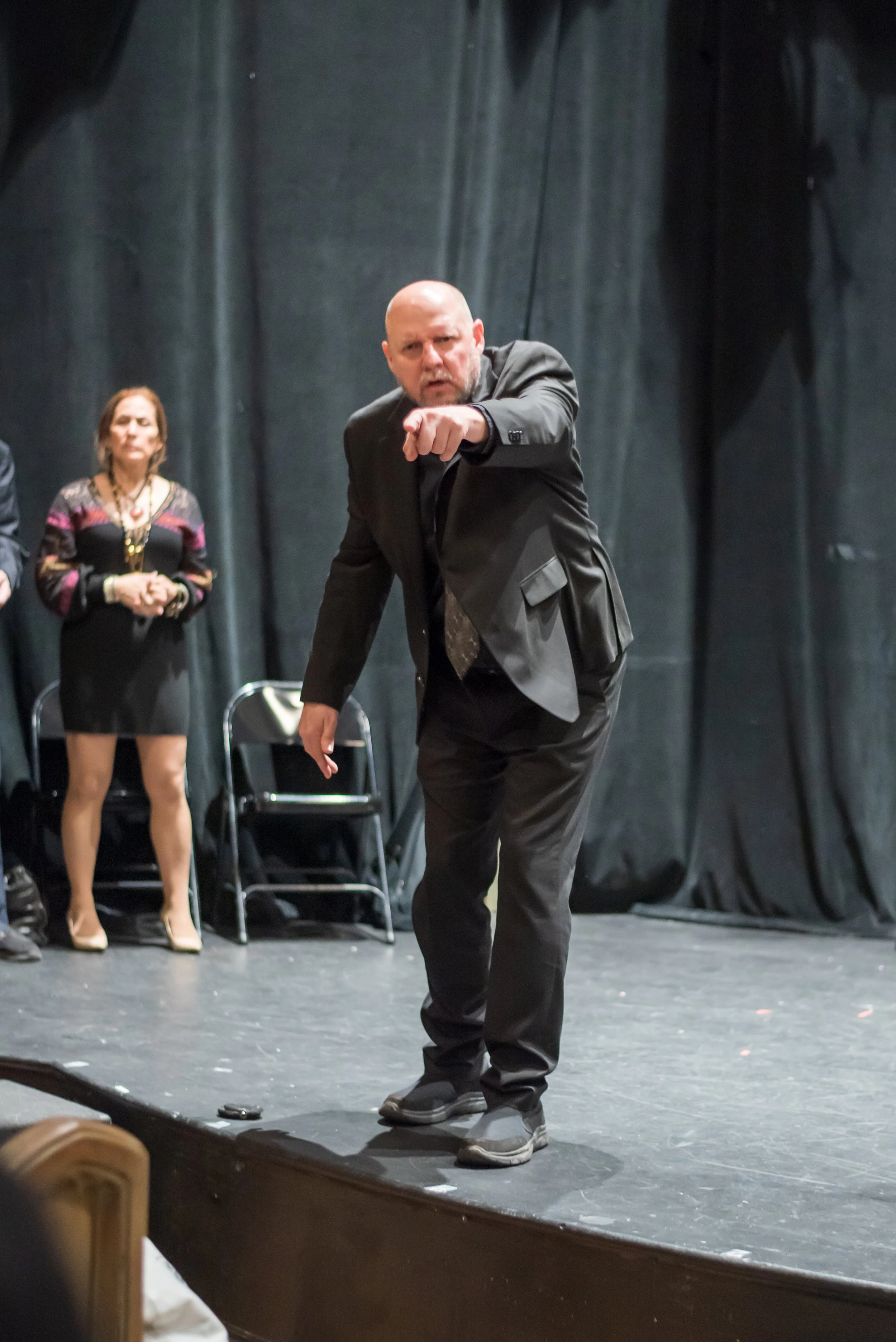 A man in a black suit pointing forward on a stage with black curtains behind him, with a woman in a black dress and patterned top and a necklace standing in the background.