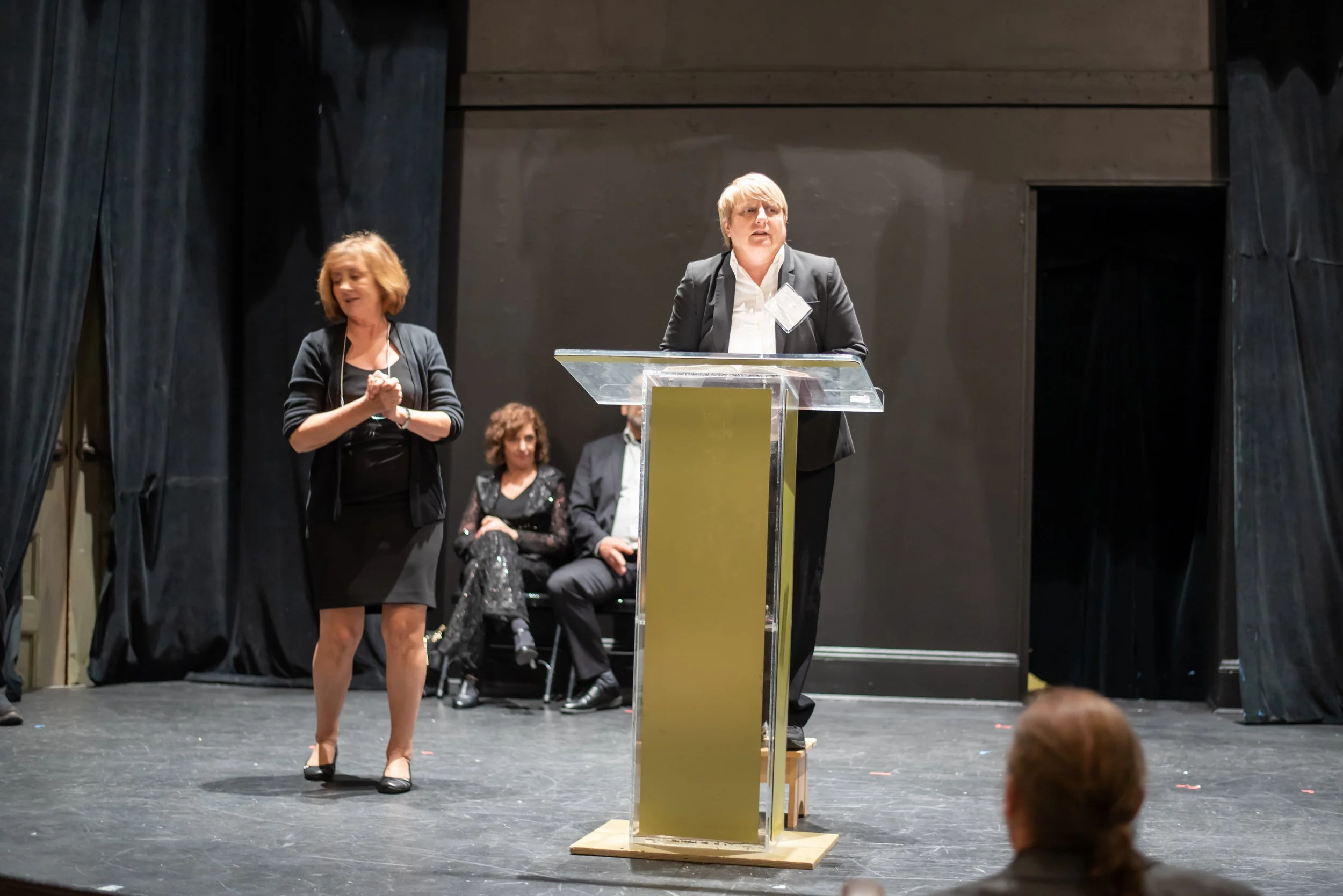 A woman is giving a speech at a podium on a stage, with three seated individuals in the background and an audience member in the foreground.