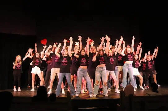 Group of women performing a dance routine on stage, with hands raised, wearing matching black shirts and jeans, some holding red hearts.
