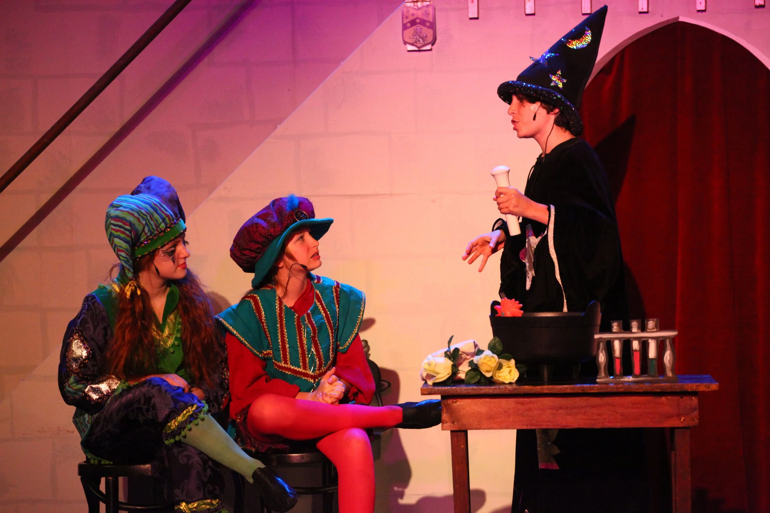 Three women dressed in colorful costumes sitting on chairs, facing a woman dressed as a witch in a black robe and pointed hat who is holding a scroll, with flowers on a table, in a theater production of Once Upon a Mattress in 2011.