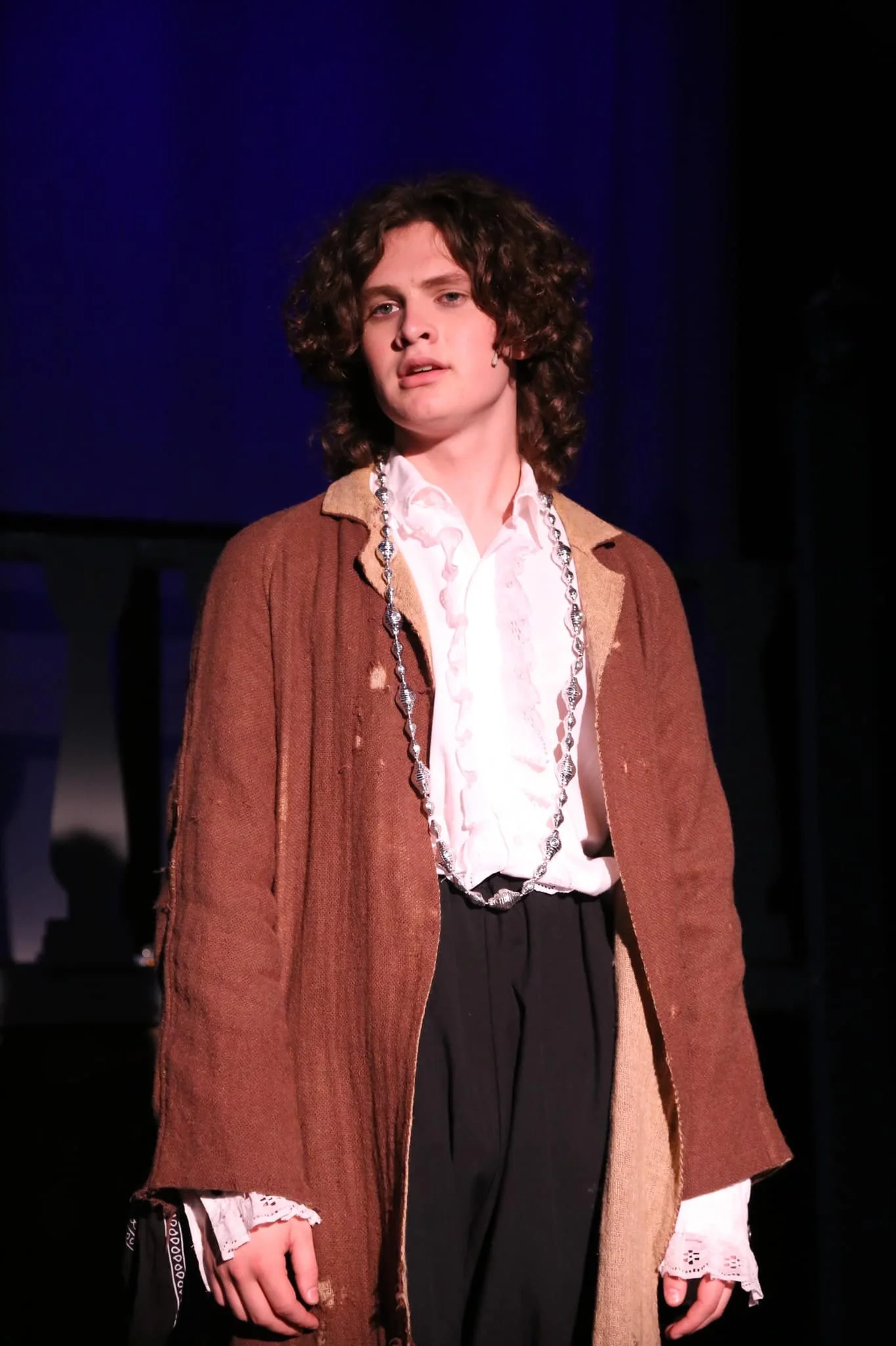 Young person with curly hair wearing a brown vintage coat, white ruffled shirt, and a long beaded necklace on stage.