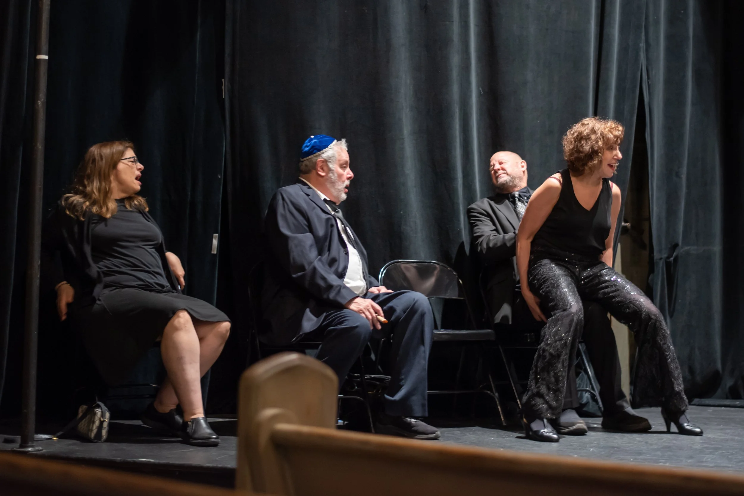 Four performers on stage, three are sitting and one is standing. They are engaged in a theatrical performance of Grandma Sylvia's Funeral with black curtains as the backdrop.