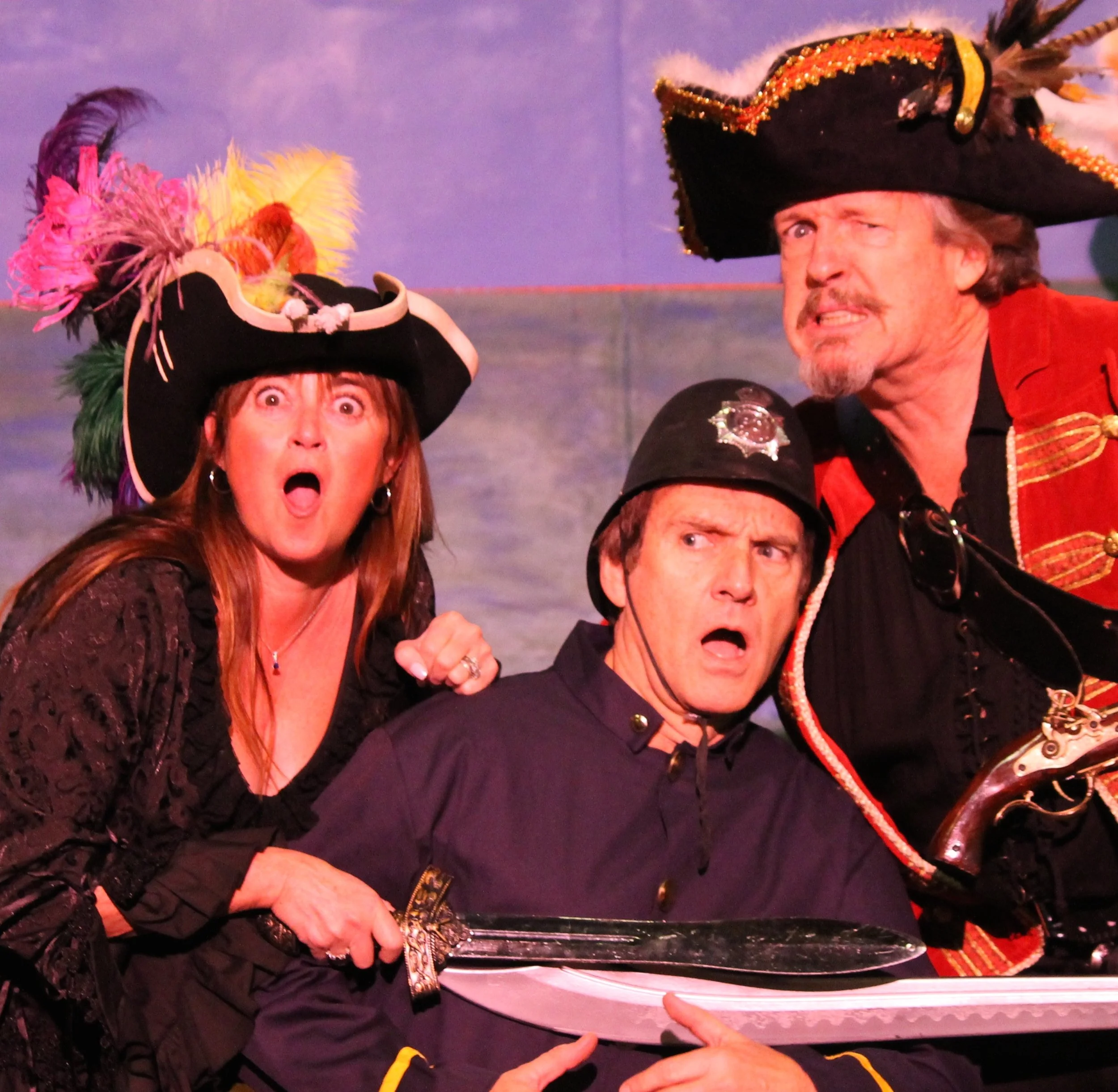 Three people in costumes portraying pirates and police with surprised and worried expressions, on a stage with a cloudy sky backdrop.