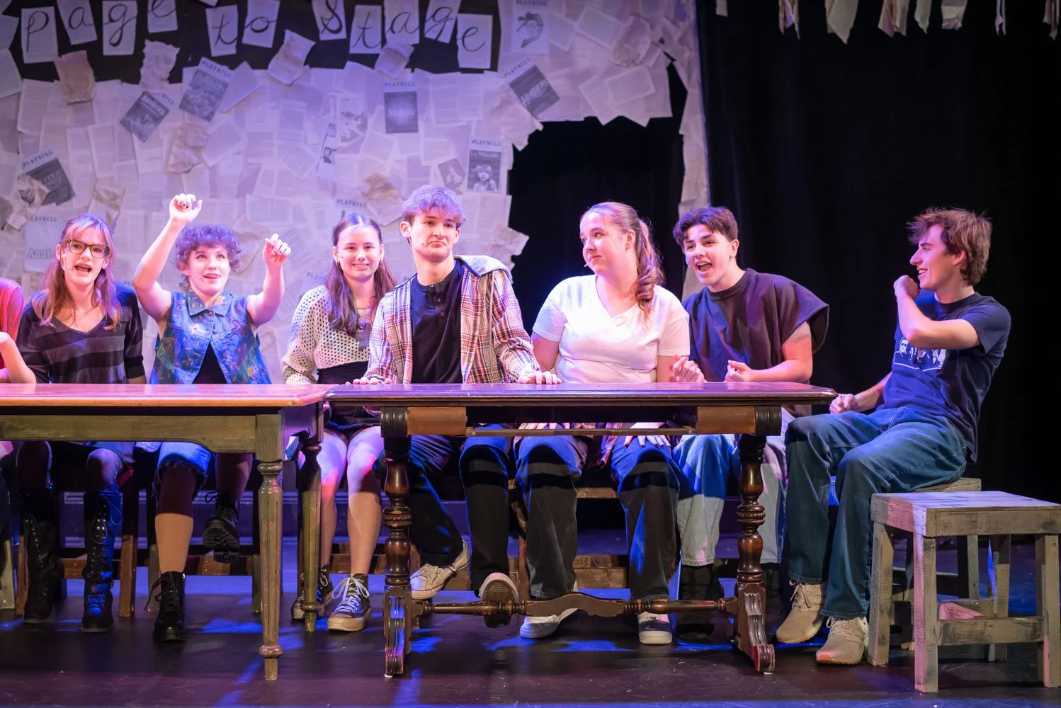 A group of seven young people sitting at a long table on stage, performing in a play with a backdrop decorated with newspapers and paper letters. Performance of Page to Stage cabaret.