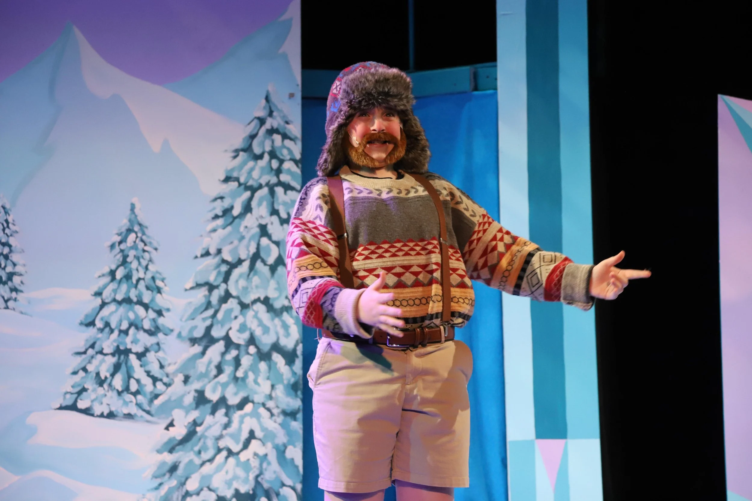 A person with a beard and mustache wearing a colorful patterned sweater, beige shorts, a furry winter hat, and suspenders, smiling and gesturing with both hands, in front of a winter-themed painted backdrop with snow-covered trees and mountains.