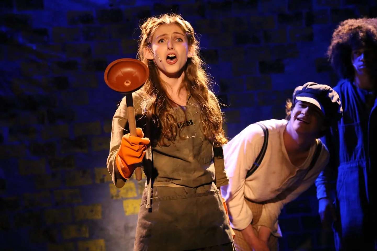 Young woman with long hair, wearing a costume and orange gloves, on stage with shocked expression, holding a brown plunger, with two other performers in the background under stage lighting.