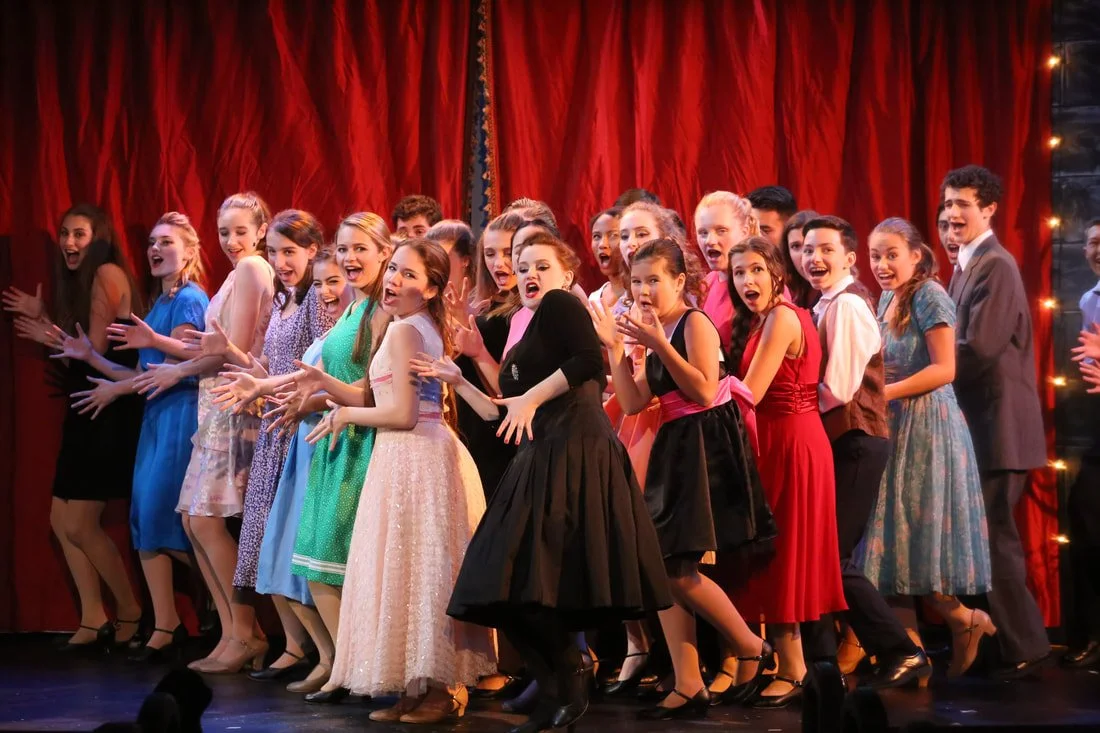 Group of children dressed in formal attire performing on stage with red curtains and string lights in the background.