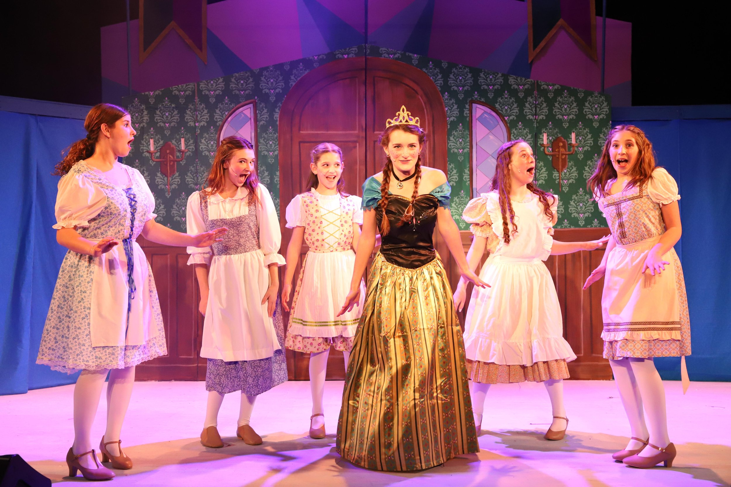 A theatrical stage scene with 7 girls in colorful costumes performing. Princess Anna, in the center, wears a tiara and a black and gold dress. The background features a painted castle interior with a large wooden door and decorative wall sconces.