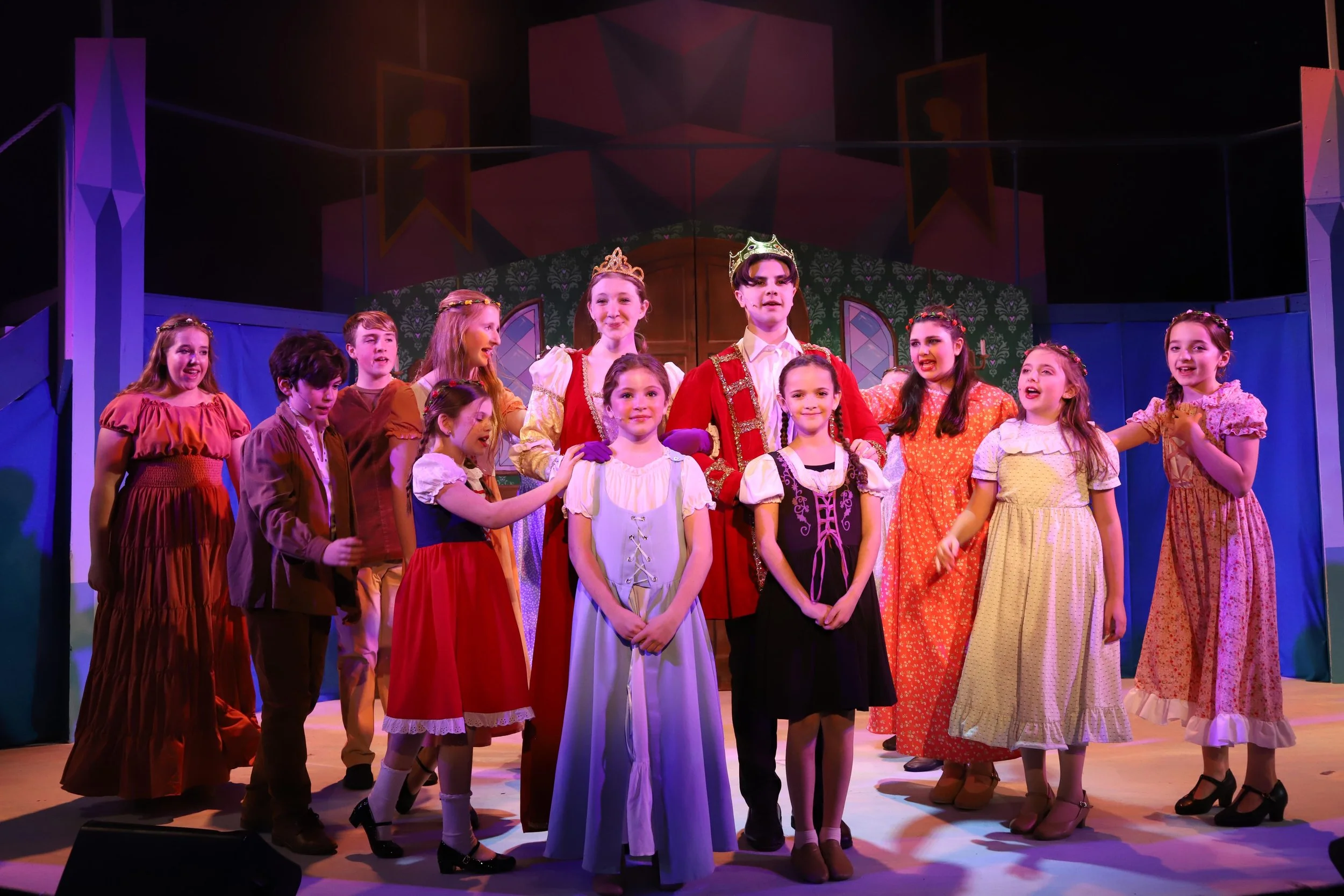 Children in colorful costumes on stage during a performance, with two children wearing crowns, suggesting they are playing royal characters in a theatrical production.