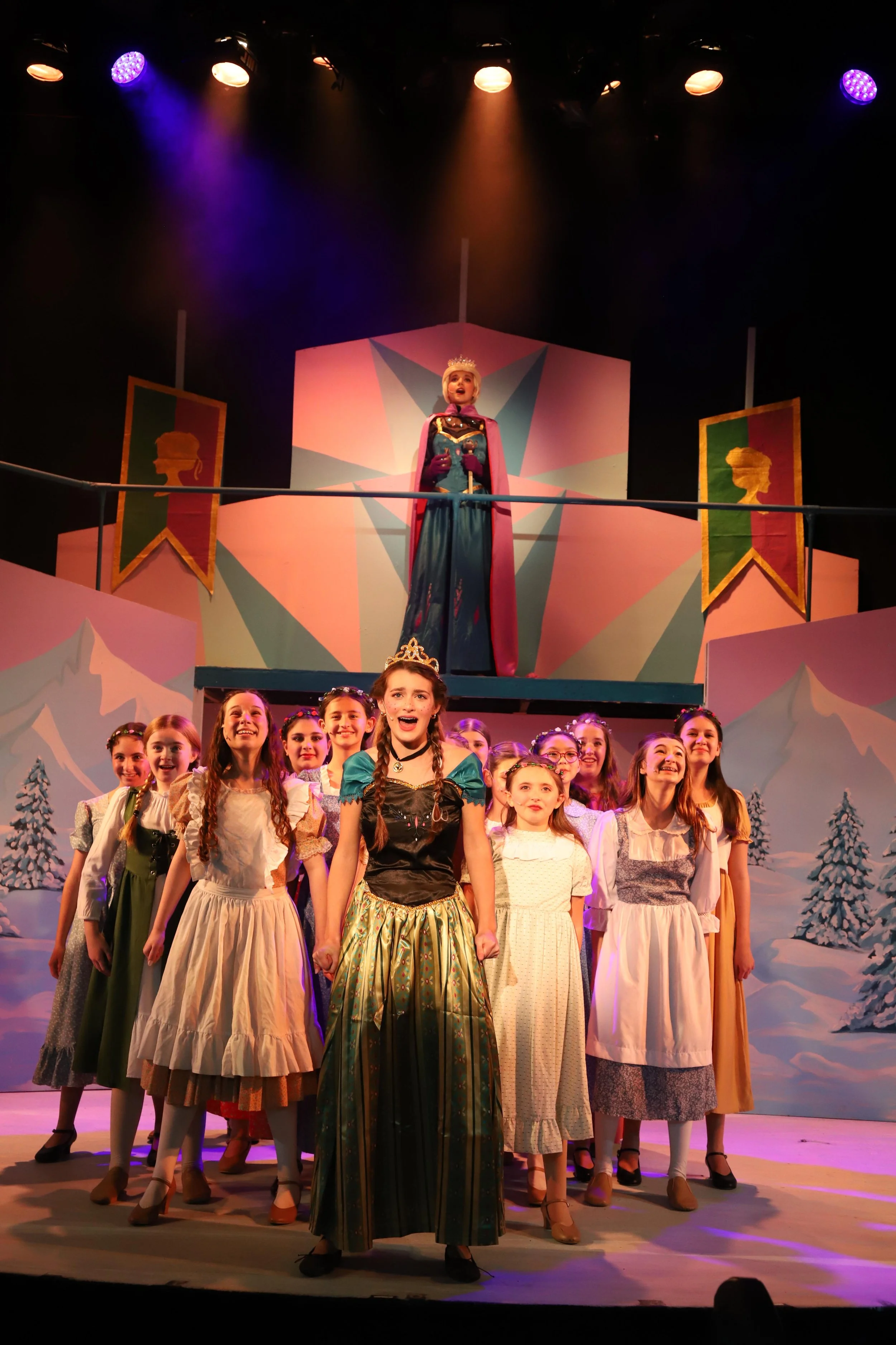Theatrical stage scene with young girls dressed as princesses, snow-covered trees in the background, and a woman dressed as a queen or princess on an elevated platform.