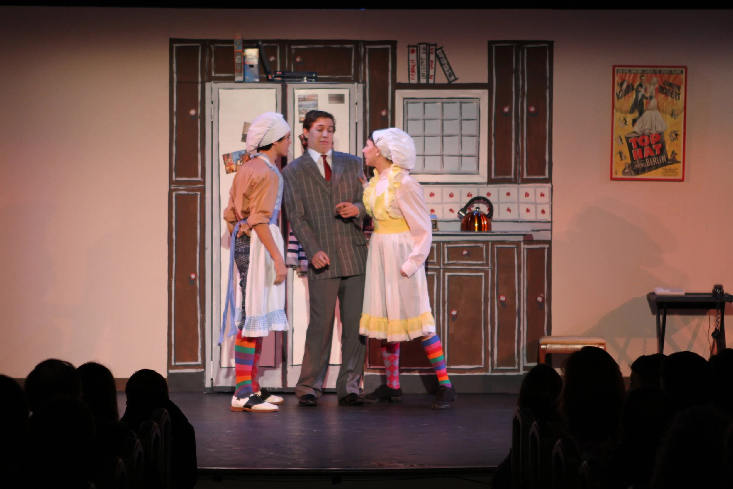 Three actors on stage performing in front of a painted kitchen backdrop. Two are dressed as chefs with tall white hats and colorful striped socks, while the man in the middle is in a suit. Scene from Drowsy Chaperone.