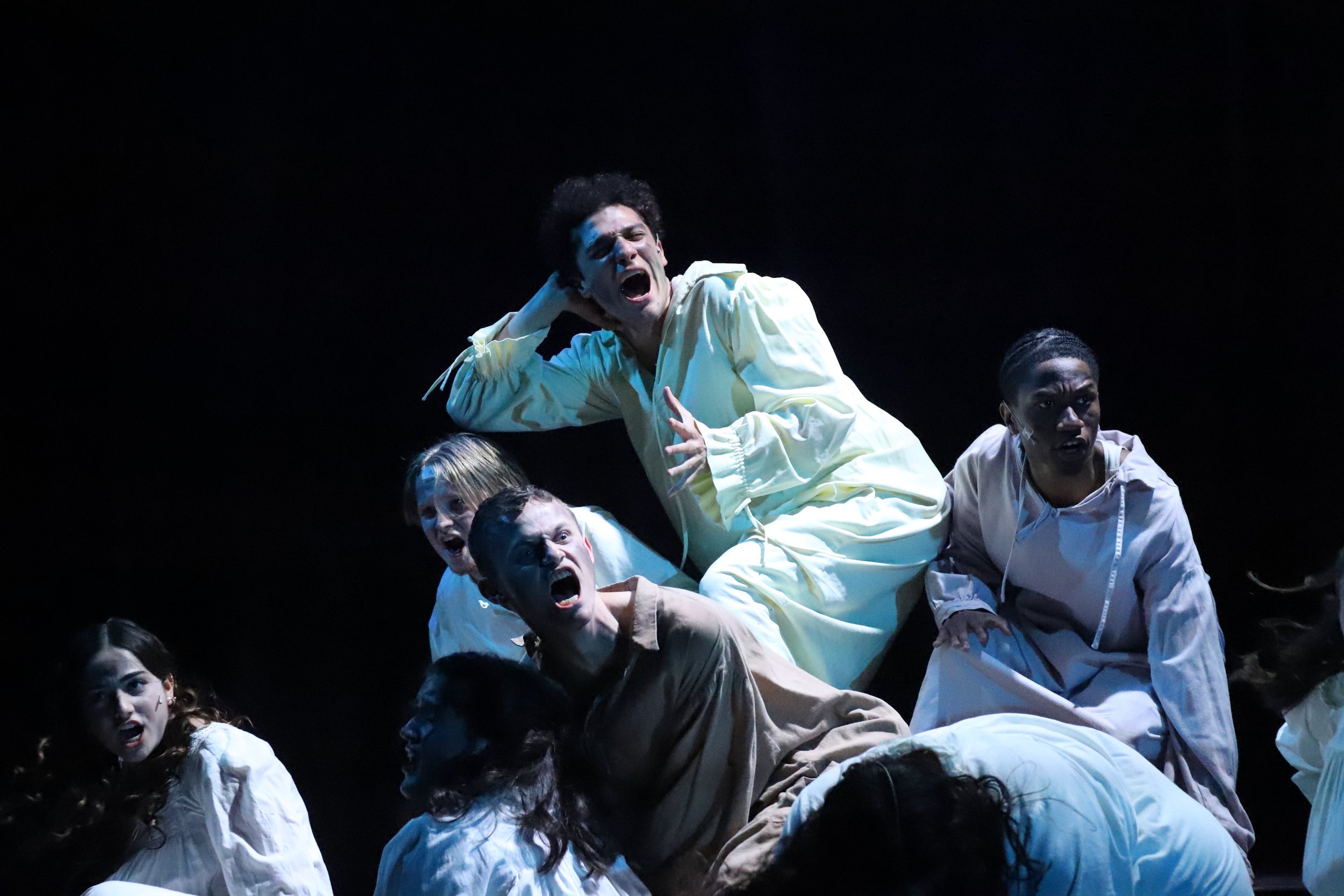 Theatrical performance with actors on stage, displaying intense expressions and gestures, dressed in light-colored costumes.