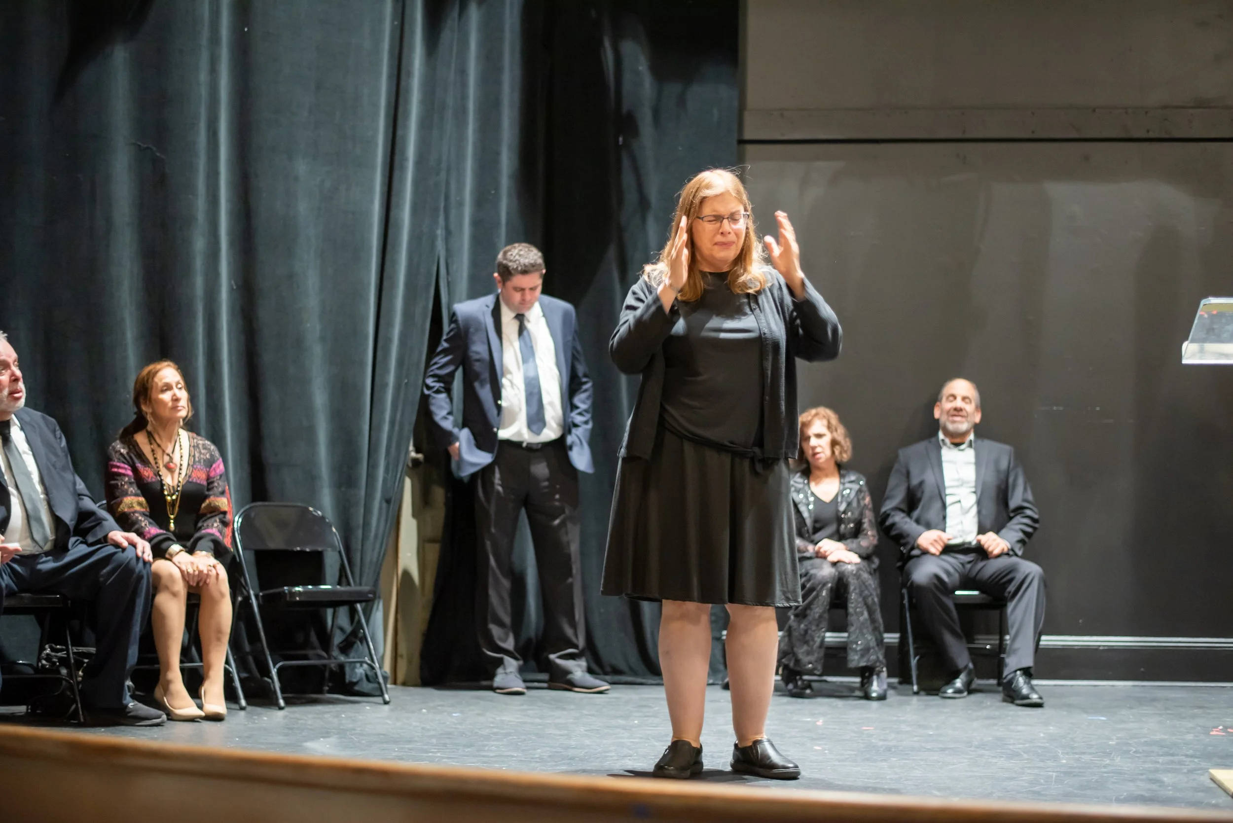 A woman in black clothing stands on a stage, with her hands near her head, appearing to be in mid-performance or speech. Behind her, five seated individuals in formal attire watch her, with a dark curtain backdrop.