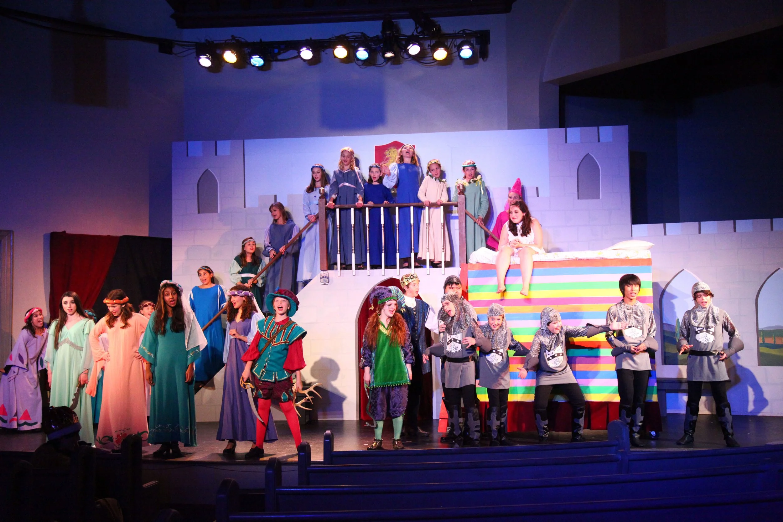Children performing in a school play depicting a scene from Once Upon a Mattress, with some dressed as princesses and others as characters in a castle setting.