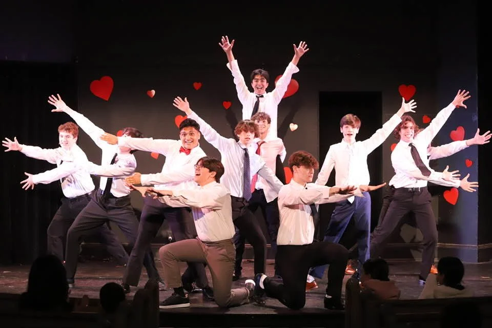 A group of young people performing a choreographed dance on stage, with a black background decorated with red and pink hearts, dressed in white shirts and dark pants.