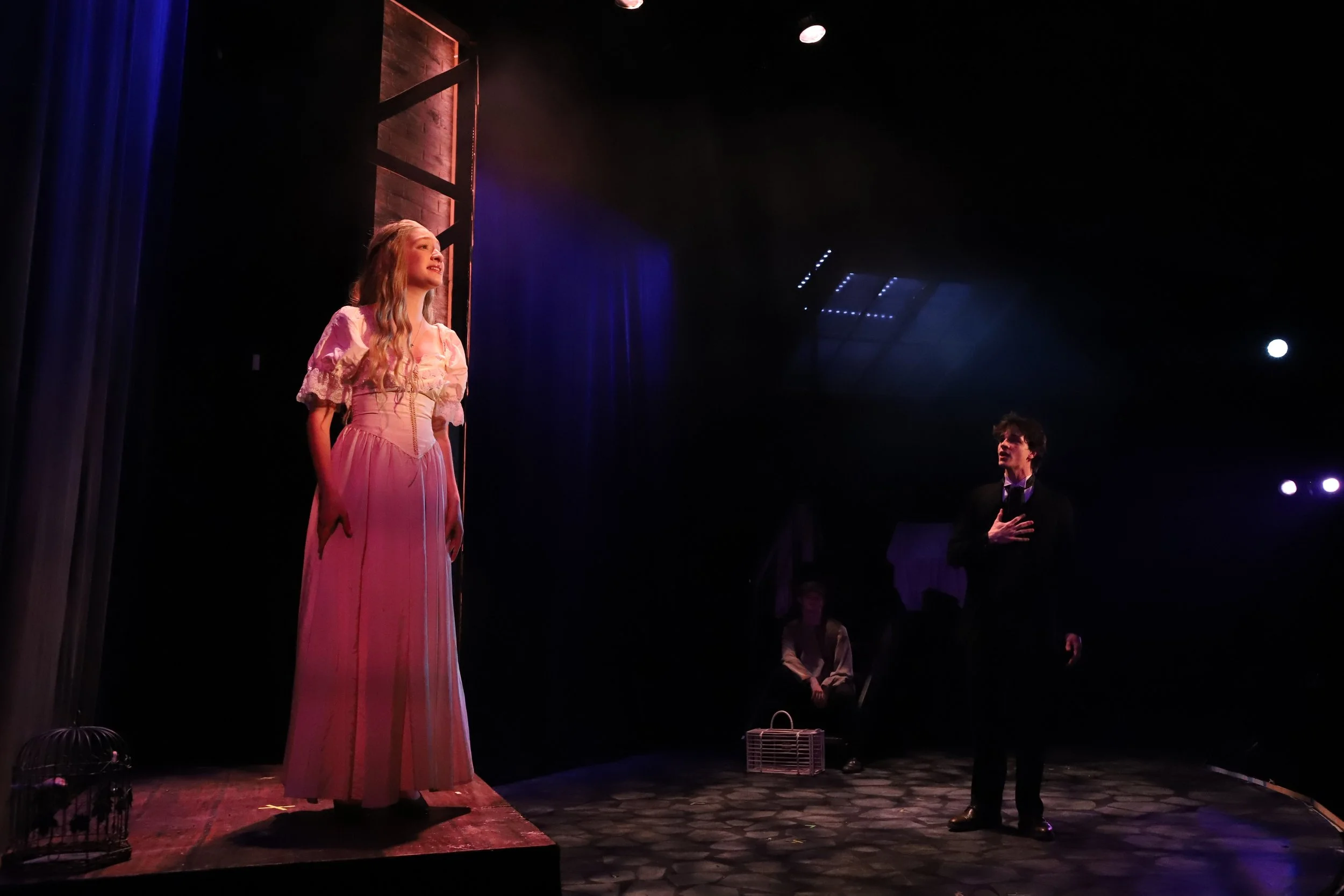 A theatrical performance with a woman in a pink dress standing near a wooden window frame on the stage, a man dressed in black with his hand on his chest, and a person sitting in the background. The stage is dimly lit with spotlight effects.